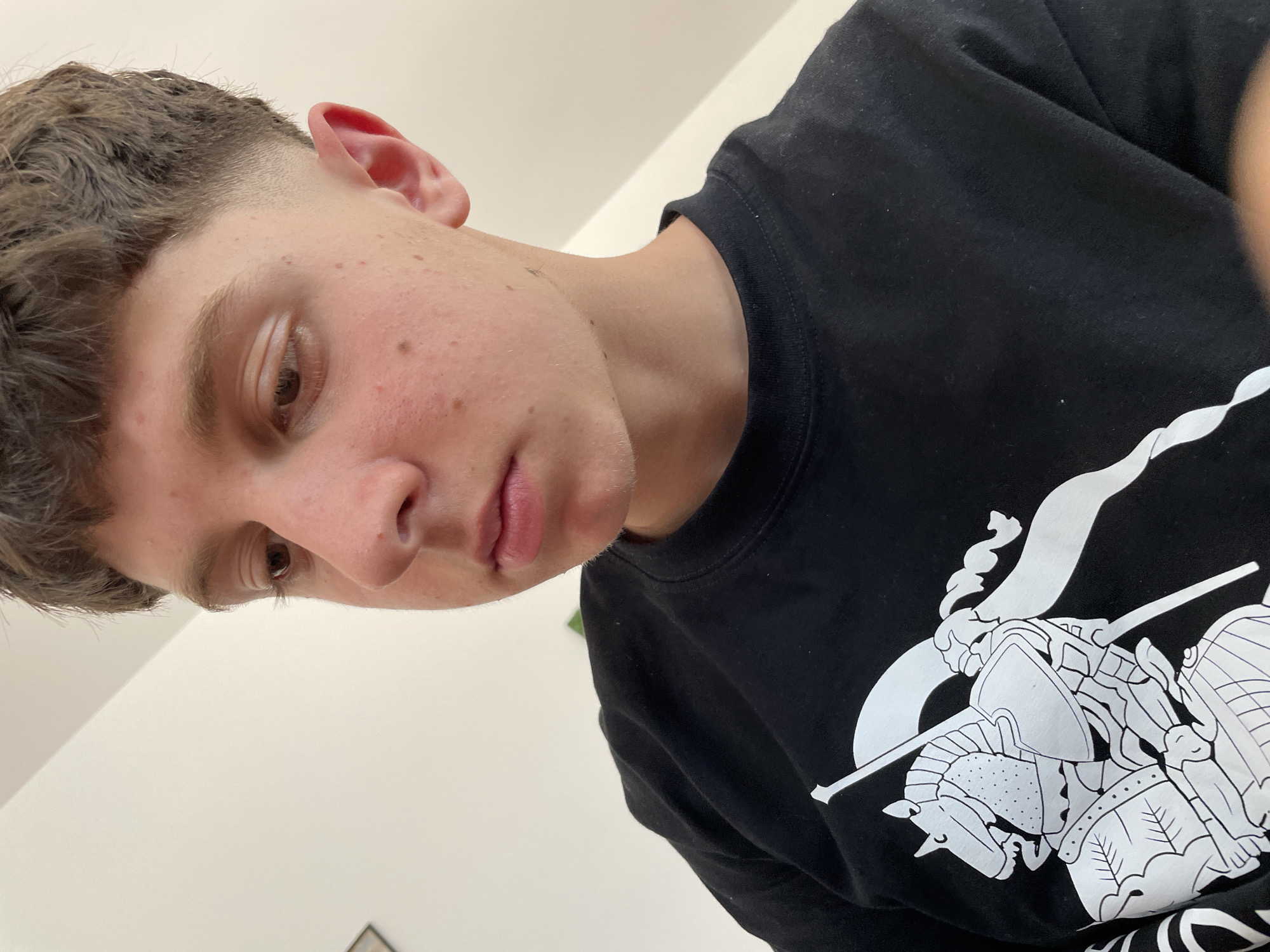 A close-up, head-and-shoulders shot shows a young person of East European appearance, likely male, with short, choppy brown hair and a visible fade haircut. The person is looking down and to the right, with their face slightly angled away from the camera. Their skin is fair with some blemishes. They are wearing a black crew-neck sweatshirt with a large, white graphic of a knight on horseback in a stylized, outline design. The background is a plain, off-white wall, suggesting an indoor setting, possibly a home. There is no indication of the specific location within Zlatograd, Bulgaria.