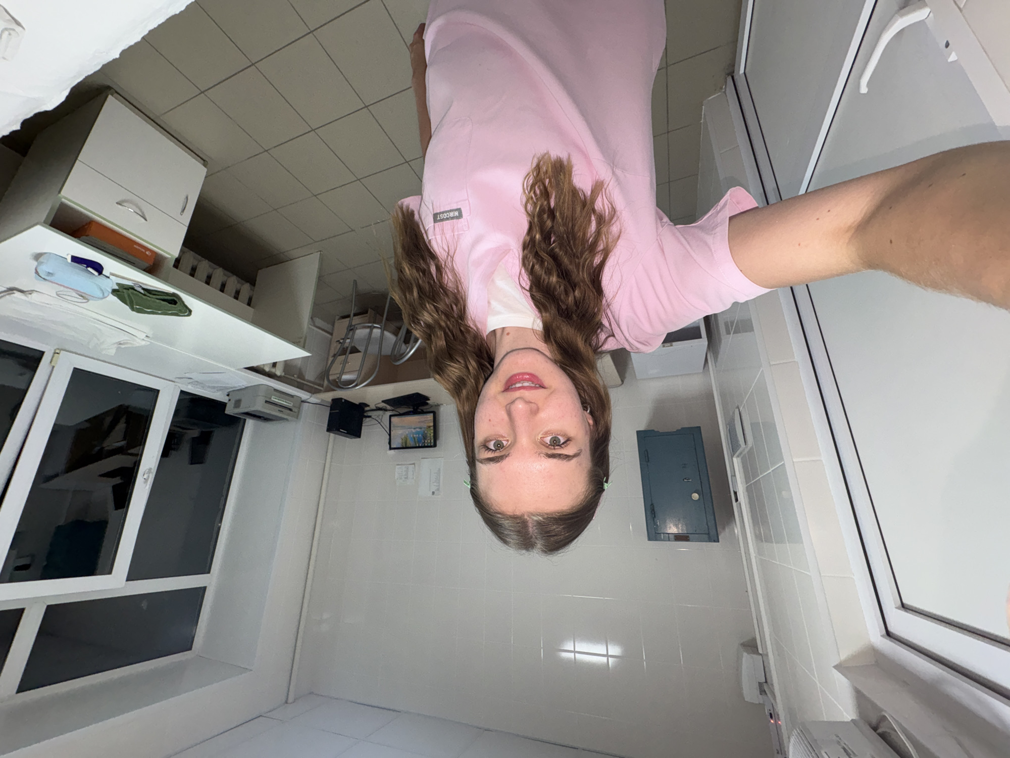 A young Caucasian woman with long, wavy brown hair, wearing a pink medical scrub top over a white t-shirt, is taking a selfie in a room that appears to be a medical office or clinic in Perm, Russia. She is standing and facing the camera, holding it with her left arm outstretched. The room has white tiled walls and a tiled floor. To her left, there is a white door with a silver handle. On the wall behind her, there is a teal metal cabinet. In the background, there is a desk with a computer monitor, keyboard, and printer. A metal chair is positioned at the desk. To the right of the desk, there is a large window with tinted panes. A white cabinet with drawers is located beneath the desk. Several items, including a green cloth, a blue roll, and a white cloth, are on the desk. The overall impression is that of a functional, perhaps slightly dated, medical workspace.