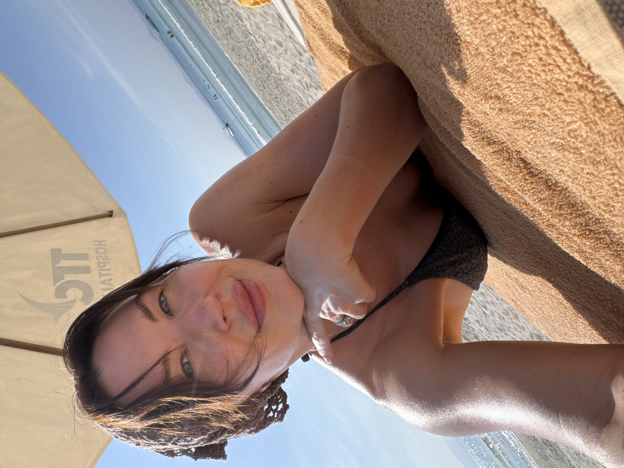 A woman is depicted lying on her stomach on an orange terry cloth beach towel spread across light-colored sand on a beach in Dong Ninh Hoa Ward, Vietnam. She rests her chin on her right hand, which wears a silver ring, and looks directly at the camera with a slight smile. Her brown hair is partially covered by a dark woven headscarf, and she has light eyes and visible freckles on her face. She is wearing a dark, possibly sparkling, bikini top. Overhead, a beige beach umbrella with the mirrored text "TTC HOSPITALITY" is partially visible. In the background, a clear blue sky transitions to a calm ocean with gentle white waves breaking on the shore. Multiple small boats are present in the distant water, with a faint landmass visible on the horizon. The scene is brightly illuminated by sunlight.