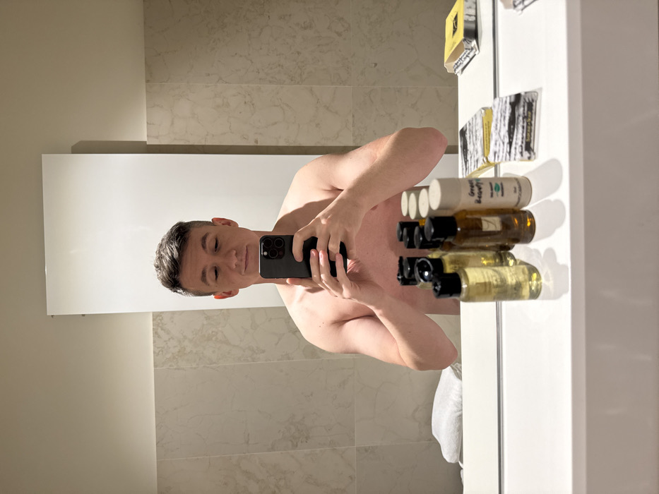 A male individual is depicted in a mirror selfie, shirtless from the chest up, within an indoor environment. He possesses short, dark hair and is observed looking downwards at a dark-colored smartphone, which he holds with both hands. The setting appears to be a bathroom, characterized by light-colored, veined marble or tiled walls. A prominent white countertop beneath the mirror displays an array of toiletries: multiple small bottles with dark caps and varying liquid contents, several rectangular packaged items (some with black and white patterns, others solid yellow), and a folded white towel. One of the cylindrical amenity bottles is labeled "Great Beauty - The Organic Skincare Collection," and another bottle's label partially shows "VENEZIA," indicating the location is in Venice, Italy. A tall, slender white panel is visible mounted on the wall behind the reflected person.