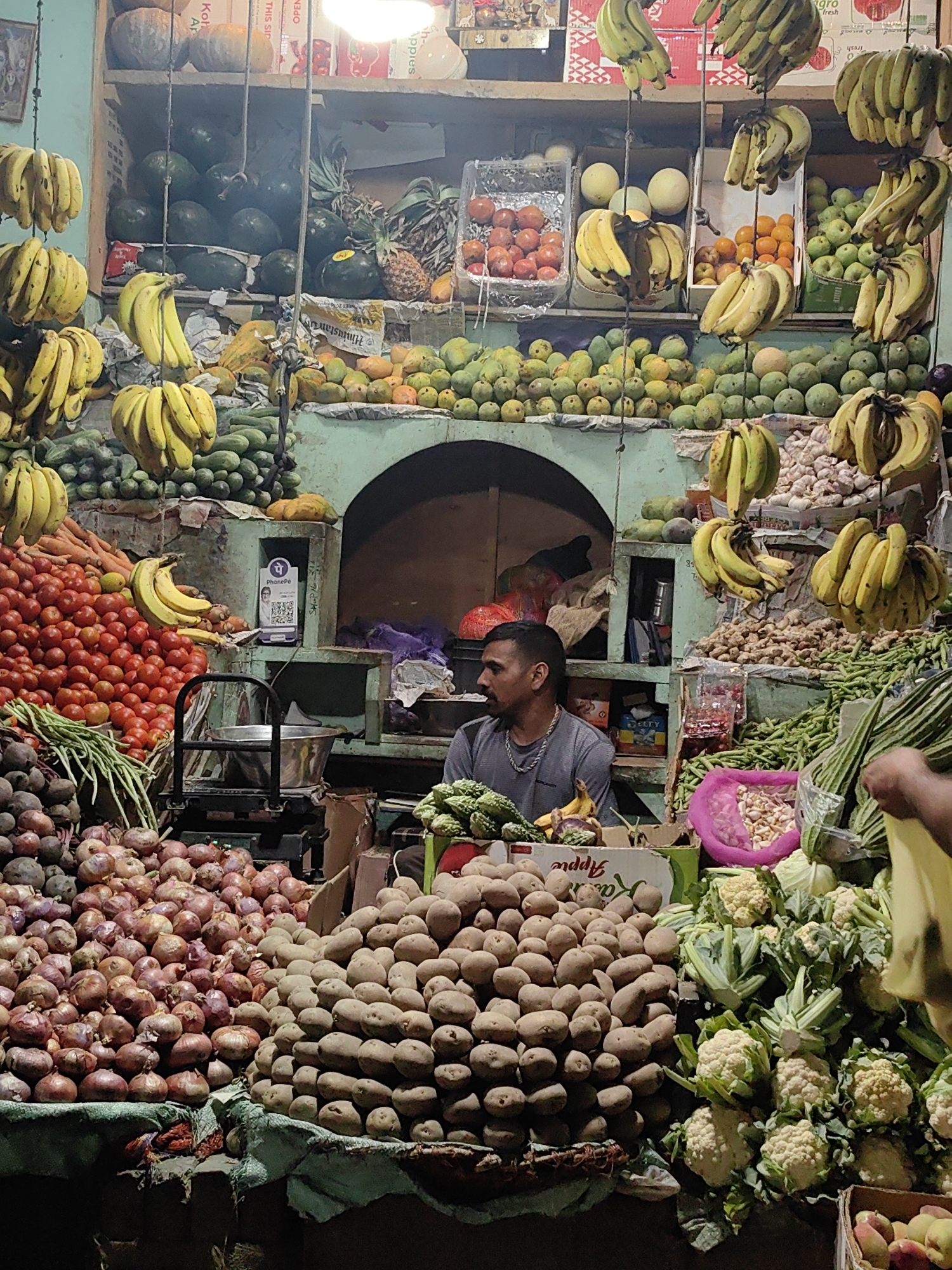 The image depicts a vibrant fruit and vegetable market stall, likely in Manali, India, given the presence of bananas, pineapples, and other produce commonly found in such regions. The stall is densely packed with a colorful assortment of fresh goods. In the foreground, large mounds of red onions and potatoes are prominently displayed. To the right, cauliflower and cabbage add to the verdant display, while a crate of what appears to be apples is visible at the very bottom right.
Several bunches of ripe bananas hang from the ceiling, creating a visual arch over the scene. Other fruits like tomatoes and what could be mangoes or other tropical fruits are visible in baskets and on shelves. The backdrop features shelves stocked with more produce, including what look like watermelons or gourds.
A man, likely the vendor, is partially visible in the mid-ground, seated and looking to his left. He is wearing a grey t-shirt and a chain necklace. The stall itself appears to be made of wood and metal, with a rustic, open-air feel. Signs and price tags are interspersed among the produce. A digital payment QR code sticker, possibly for a service like PhonePe, is affixed to a stand. The lighting is somewhat dim, suggesting an evening or indoor setting, but the produce is well-lit. The overall impression is one of abundance and lively commerce.