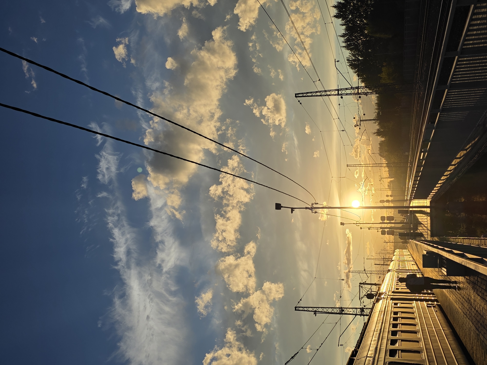 A train car, illuminated by the low-lying sun, is parked at a station platform in Posolok Kombinata Stroymaterialov-2, Russia. A lone figure, silhouetted against the vibrant sky, walks away from the camera down the platform. Overhead, power lines crisscross the frame, juxtaposed against a dramatic sky filled with scattered clouds catching the golden light of sunset. In the distance, trees line the horizon, and the glow of the setting sun casts long shadows across the tracks.