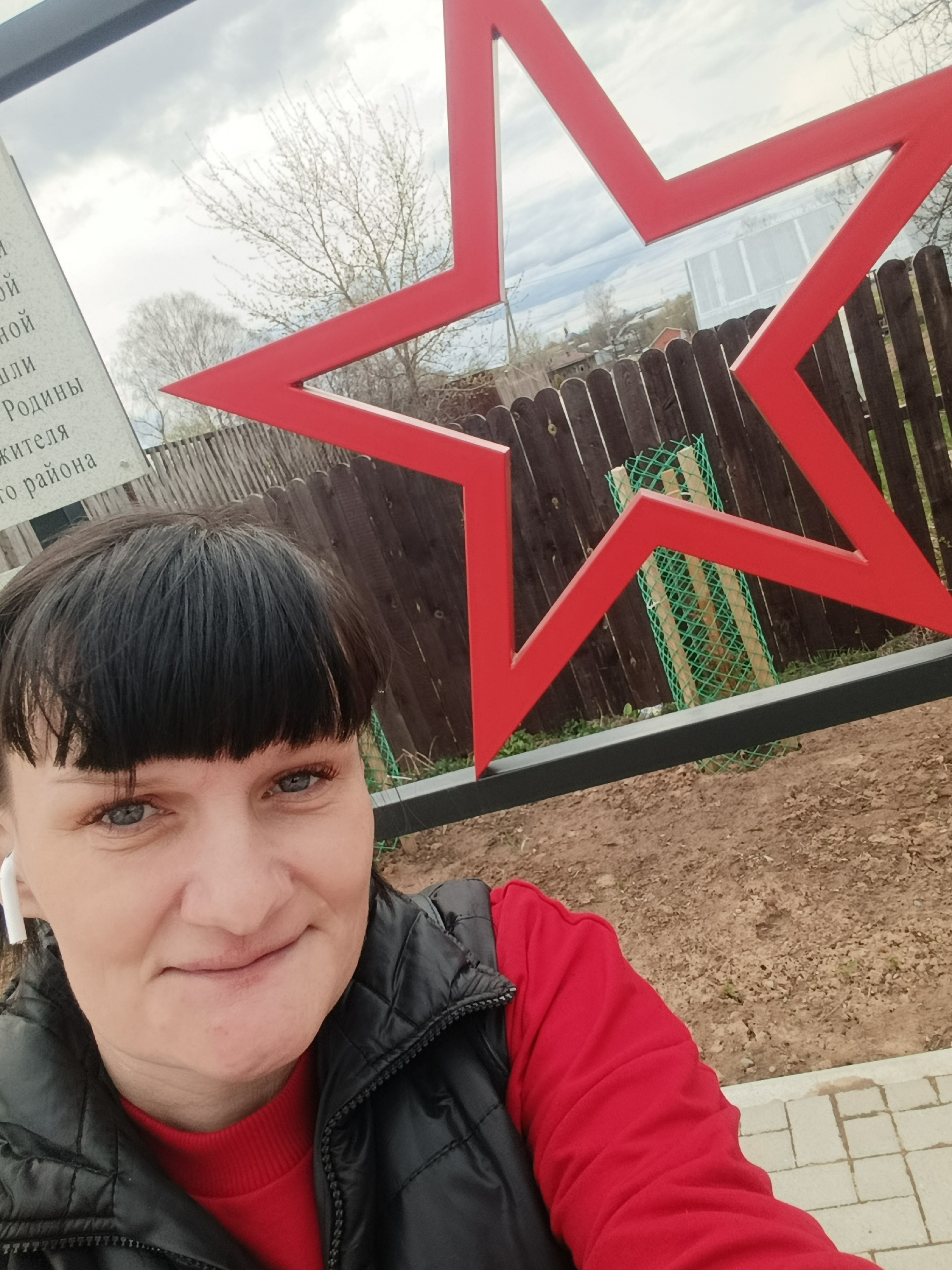 A woman is captured in a selfie-style shot, smiling faintly at the camera. She has dark hair with bangs, blue eyes, and is wearing a black puffer vest over a red long-sleeved top, with a white earbud visible in her left ear.
Behind her, a large, striking red five-pointed star structure dominates the background, suggesting a monument or public art installation. The star partially frames a wooden fence and sparse trees with bare branches, indicative of late winter or early spring. The ground is visible as dirt and some paving stones at the bottom right. The sky is overcast and cloudy.
To the left, a memorial plaque or sign is partially visible, with text in Cyrillic (Russian). Observable words and fragments include "...и", "ной", "ой", "ли", "Родины" (Homeland/Motherland), "жителя" (resident/inhabitant), and "о района" (of the district/region). This text, combined with the prominent red star, strongly suggests a war memorial or monument, common in Russia, specifically located in Фоки. The overall scene conveys a daytime setting with diffused light from the cloudy sky.