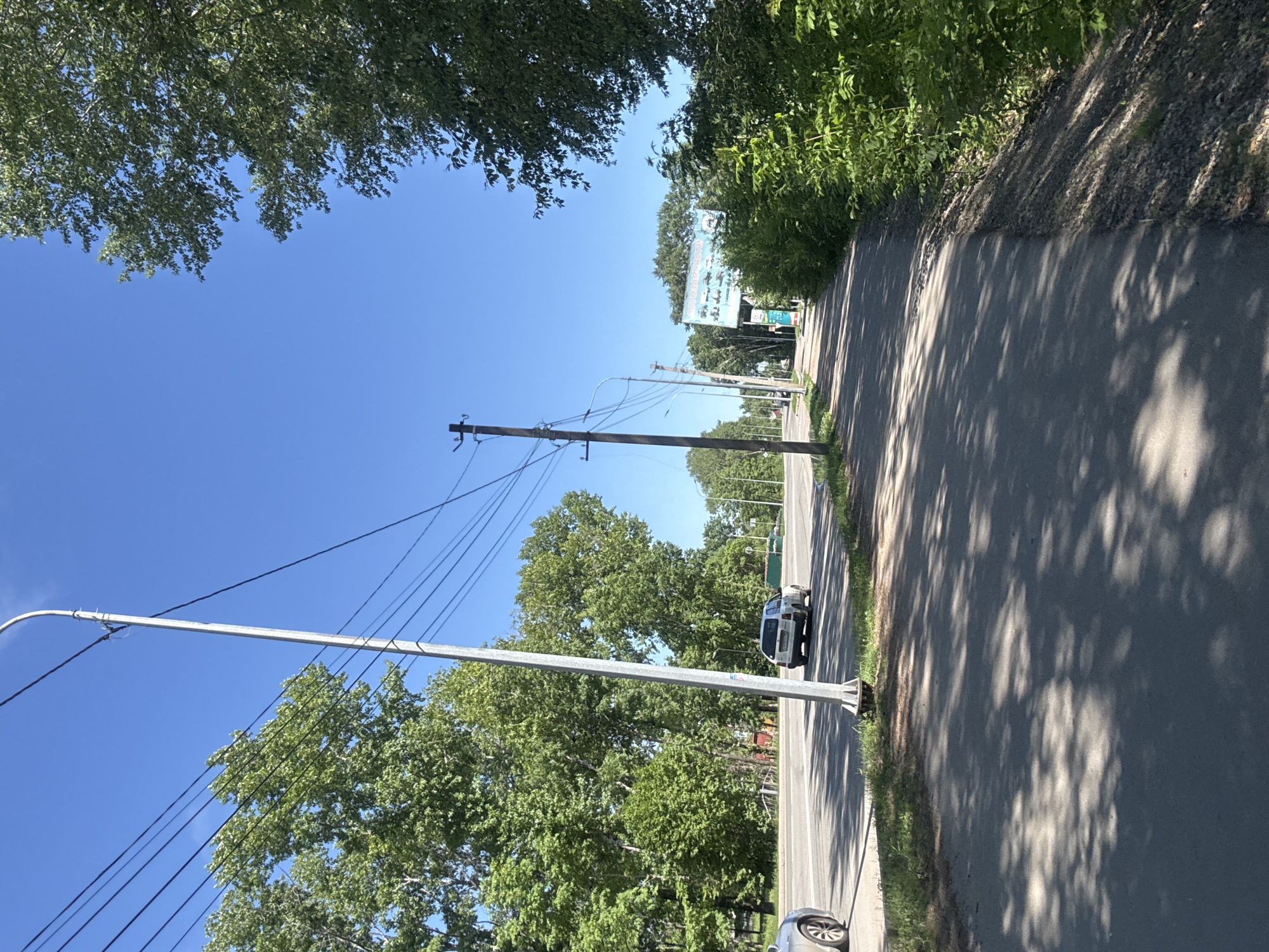The image depicts a sunny day in Birobidzhan, Russia, on an asphalt road with a pedestrian walkway to the right. Tall green trees line both sides of the road, casting dappled shadows on the walkway and road. On the left side of the road, there is a multi-lane road with two cars visible, one silver car partially visible and a white SUV driving away from the camera. A tall, silver lamppost stands prominently in the foreground on the left, with power lines running across the sky. On the right side of the road, further in the distance, a large billboard advertising agricultural machinery and equipment is visible. The sky is a clear, vibrant blue. The overall scene is one of a typical urban or suburban environment on a bright day, with elements of infrastructure, transportation, and natural vegetation.