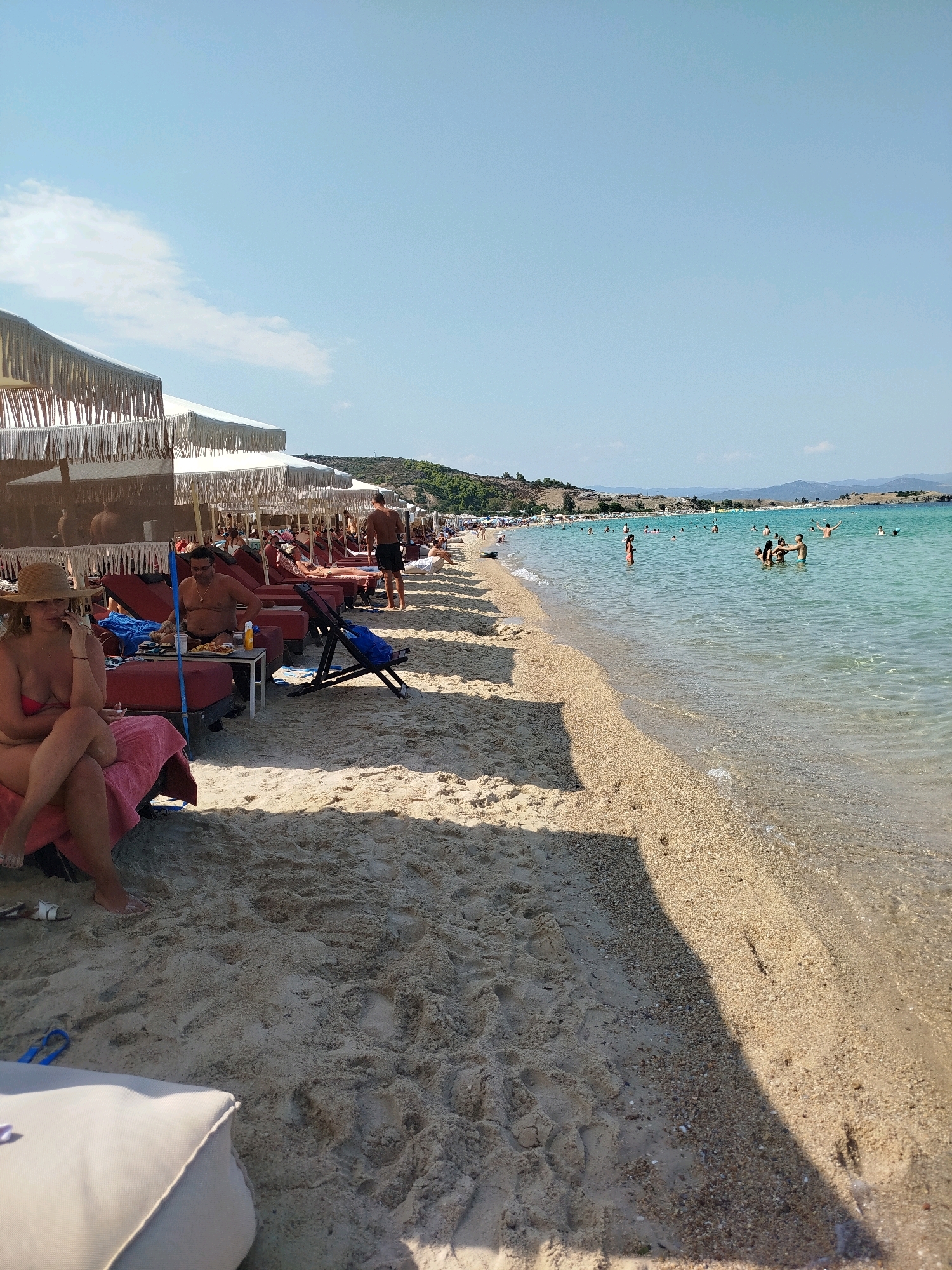 A crowded beach scene in Sithonia Municipal Unit, Greece. The image captures a sunny day with a clear blue sky. The foreground is dominated by sand, with beach chairs and umbrellas set up for relaxation. Several people are visible, some lounging on chairs, others walking or swimming in the clear turquoise water. The water is shallow near the shore and becomes deeper further out, with a few people enjoying swimming and playing in the sea. The background shows a hilly landscape with vegetation and more buildings or structures along the coastline. The overall atmosphere is one of a busy, popular beach destination.