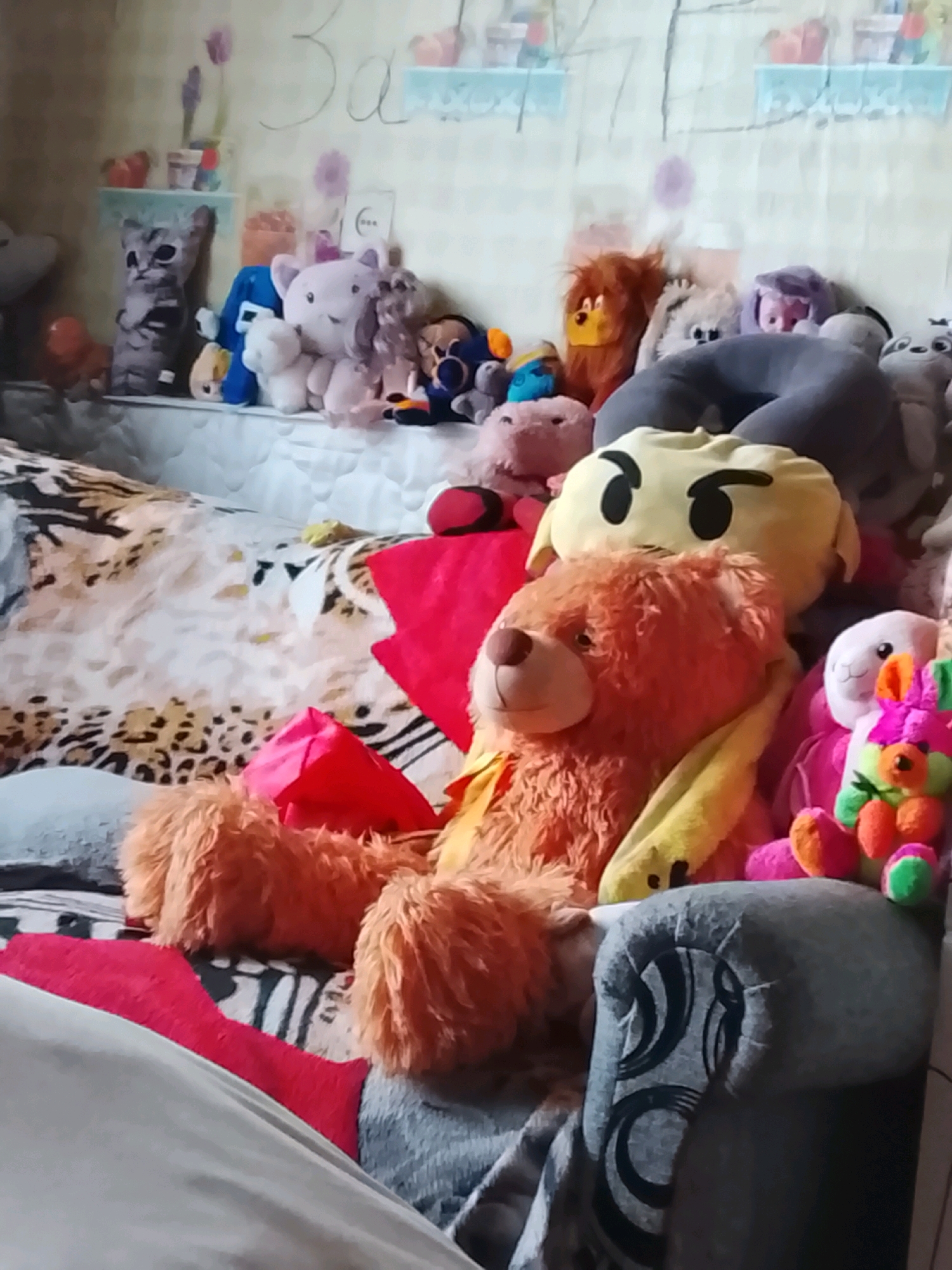 The image shows a collection of stuffed animals on a bed. The stuffed animals are of various sizes and colors, including a large orange bear in the foreground, a yellow pillow with a face, a gray cat, a pink pig, a lion, and a white rabbit. The bed appears to be covered with a patterned sheet or blanket. In the background, there is a wall with what appears to be drawings or decorations. There is no visible indication of the location being in the city of Karma, Belarus.