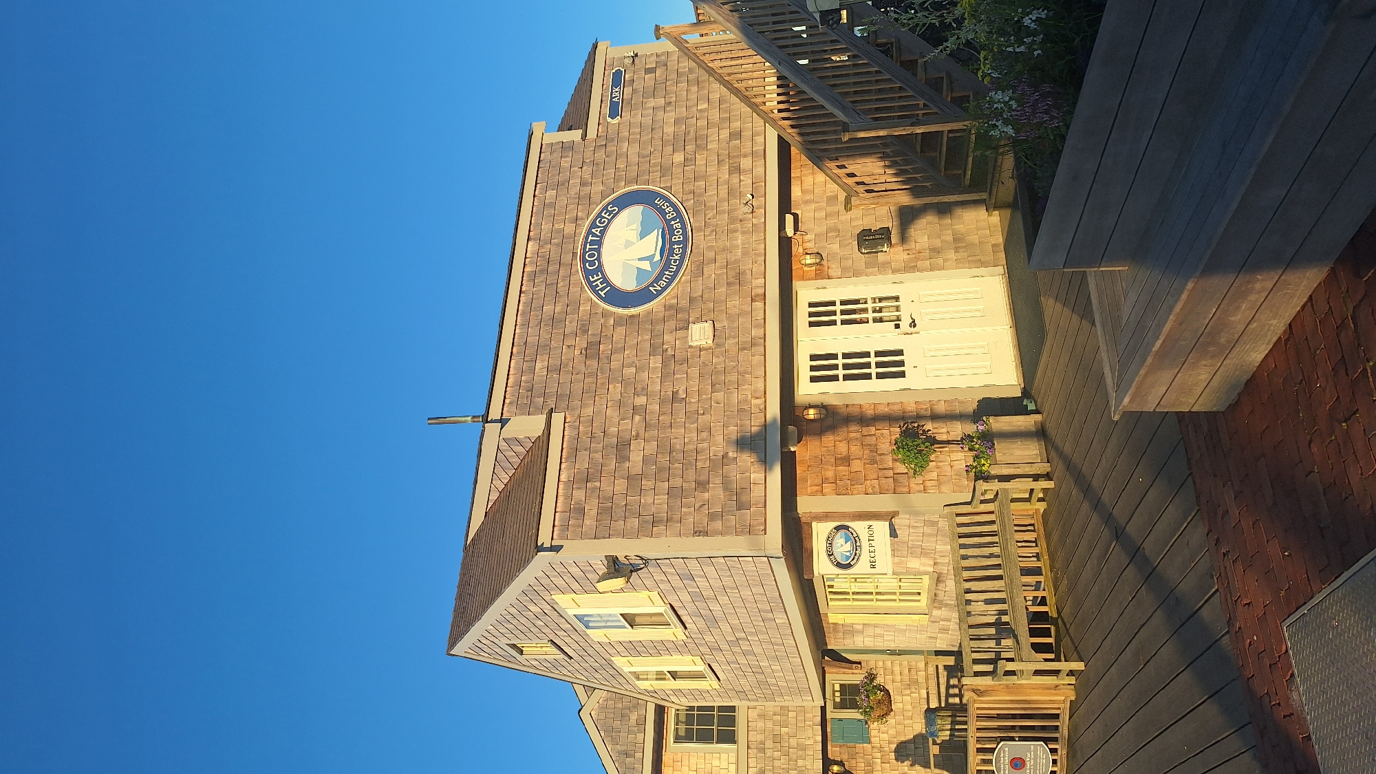 The image depicts the exterior of a building located at the Nantucket Boat Basin in Nantucket, Massachusetts, USA. The building has a distinctive shingle-style exterior and features a prominent circular sign with the text "THE COTTAGES Nantucket Boat Basin" and a sailboat graphic. Adjacent to this sign, a smaller rectangular sign reads "ARK". The building has a wooden staircase leading up to an entrance with double doors. In front of the building, there is a wooden deck area with benches, planters with flowers, and a wooden railing. The sky is clear and blue, suggesting a sunny day. The overall scene conveys a coastal and possibly resort-like atmosphere.