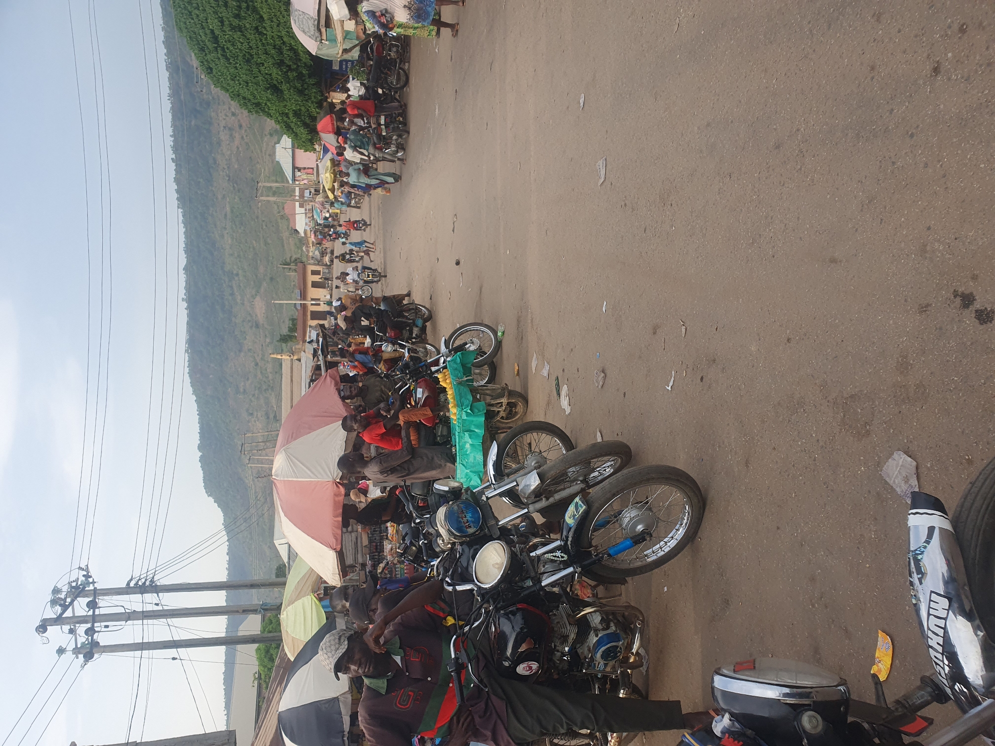 The image depicts a busy, open-air street scene in Lokoja, Nigeria, featuring a concentration of people and motorcycles alongside what appears to be a market or a local transportation hub. The perspective is slightly elevated, looking down a wide, dusty road that stretches into the distance.
In the foreground, several motorcycles, commonly known as "boda bodas," are parked. One motorcycle's chrome exhaust pipe prominently displays the word "HUNTER." A blue plastic bottle is also visible on another motorcycle. Many individuals, mostly men, are present; some are seated on their motorcycles, while others stand beside them or gather under large parasols. These umbrellas, colored pink and white, green, and blue and white, provide shade from the bright sun. One man in the foreground wears a baseball cap with a "GG" style logo. Among the parked motorcycles and people, various items like bags and what appears to be a bunch of bananas can be seen, suggesting market activities or goods being transported.
Further down the road, more people and motorcycles are visible, indicating continued activity along the street. Utility poles with multiple power lines are visible on the left side of the frame, providing a sense of the local infrastructure.
The background is dominated by a majestic, tree-covered mountain range under a clear, bright sky, characteristic of the geographical setting. The clear sky and strong sunlight suggest it is daytime. The overall scene conveys a bustling, everyday atmosphere typical of a vibrant West African community.
