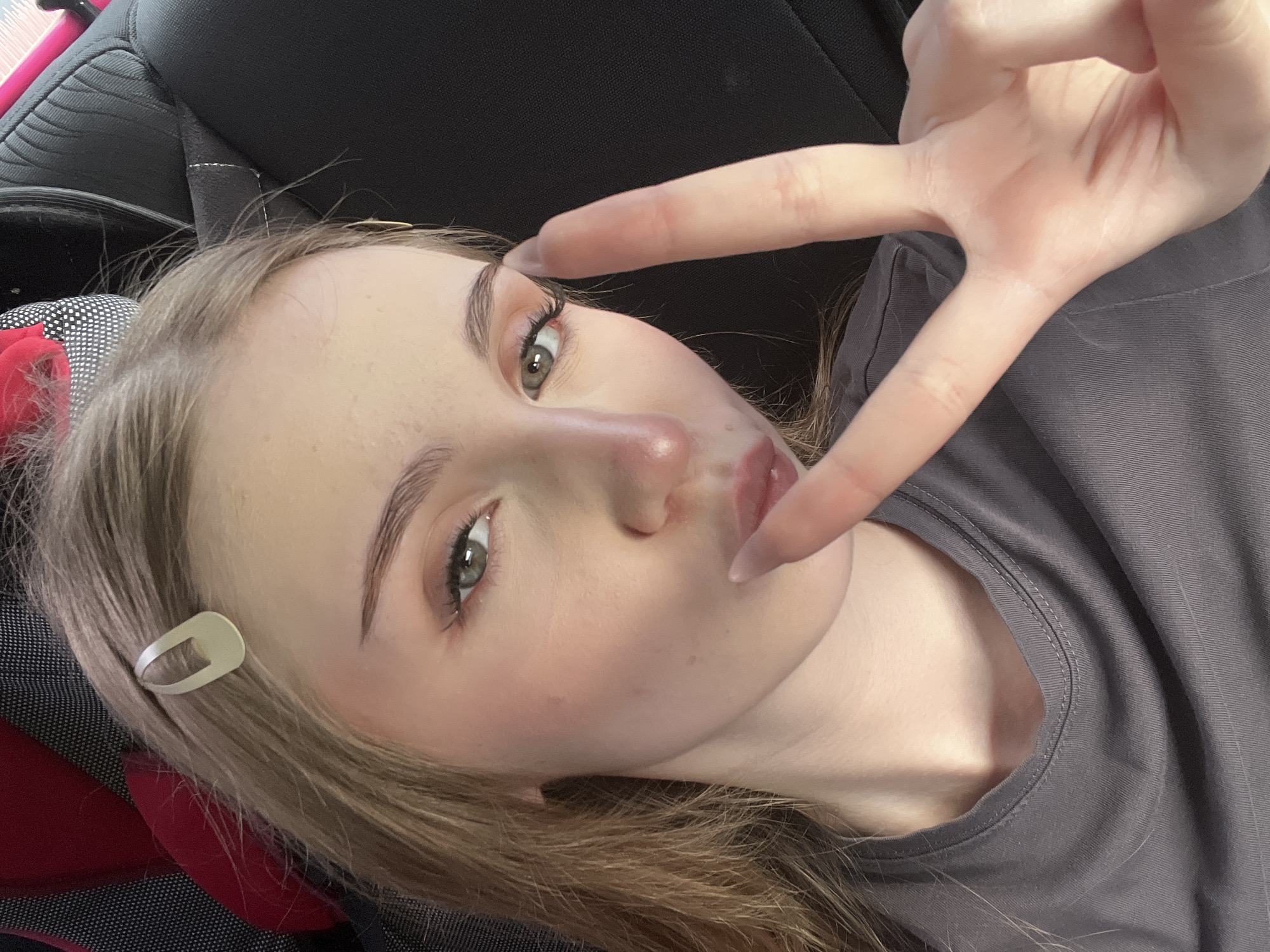 The image is a close-up, selfie-style portrait of a young woman with fair skin and blonde hair. She is wearing a grey t-shirt and has a light-colored hair clip in her hair. Her makeup includes eyeshadow and eyeliner, and her lips are slightly pursed. Her right hand is raised, forming a peace sign. The background appears to be the interior of a vehicle, with a dark, textured seat visible behind her. The lighting suggests it is daytime. There is no indication of the city of Новая Отрадовка, Russia, within the image.