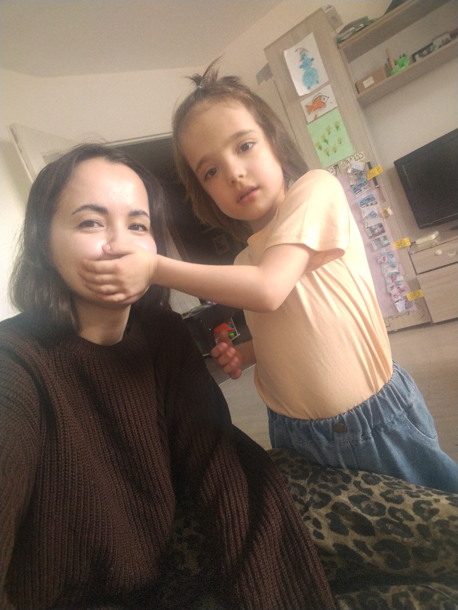 A brightly lit indoor photograph, taken in Noviy, Russia, features a woman and a young girl. The woman, with dark, shoulder-length hair, occupies the left foreground, wearing a dark brown knitted sweater. Her mouth is playfully covered by the hand of the young girl, though her visible eyes show an amused or smiling expression.
The young girl, with light brown hair tied in a small bun, stands to the right, dressed in a peach-colored t-shirt and blue elastic-waist pants. She looks directly at the camera with a neutral, perhaps curious, expression.
The background reveals a home interior, likely a living space or child's room. A light-colored wooden cabinet with shelves is visible, adorned with several children's drawings, including a blue snowman, an orange fish, and yellow flowers. A black television set is partially visible on the right. Below the shelves, a vertical strip of papers or cards is taped to a surface; discernible text includes "ДЕНЬГИ" (Money) and "СЛАВДА" (Slavda), both appearing upside down. A white label with "ТВ" is also partially visible on the cabinet. In the immediate foreground, part of a leopard print fabric, possibly a cushion or blanket, adds a textural element. The scene is illuminated by natural daylight, suggesting a daytime setting.