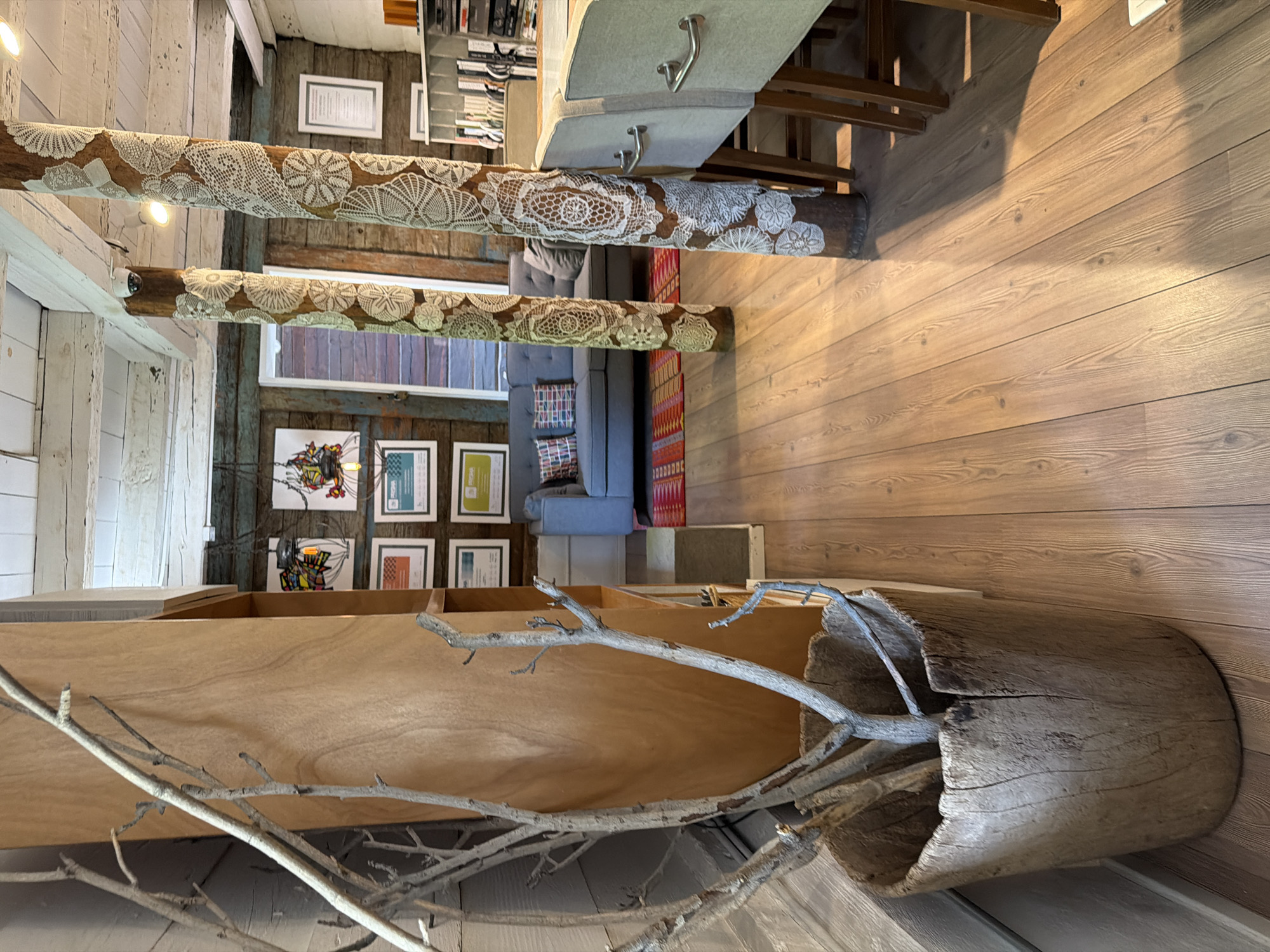 The image shows an interior space with a rustic design. The walls are a mix of wooden planks and decorative elements. Two wooden beams covered in lace-like fabric run across the upper part of the room, with embedded light fixtures. A table with chairs sits near the beams. A gray sofa and colorful rugs are visible in the background. A decorative arrangement with branches inside a hollowed-out log is located in the foreground. The flooring is made of wood planks. Framed artwork adorns the walls. Without any information about the city of Puebla, Mexico, I cannot determine if this scene is located there.