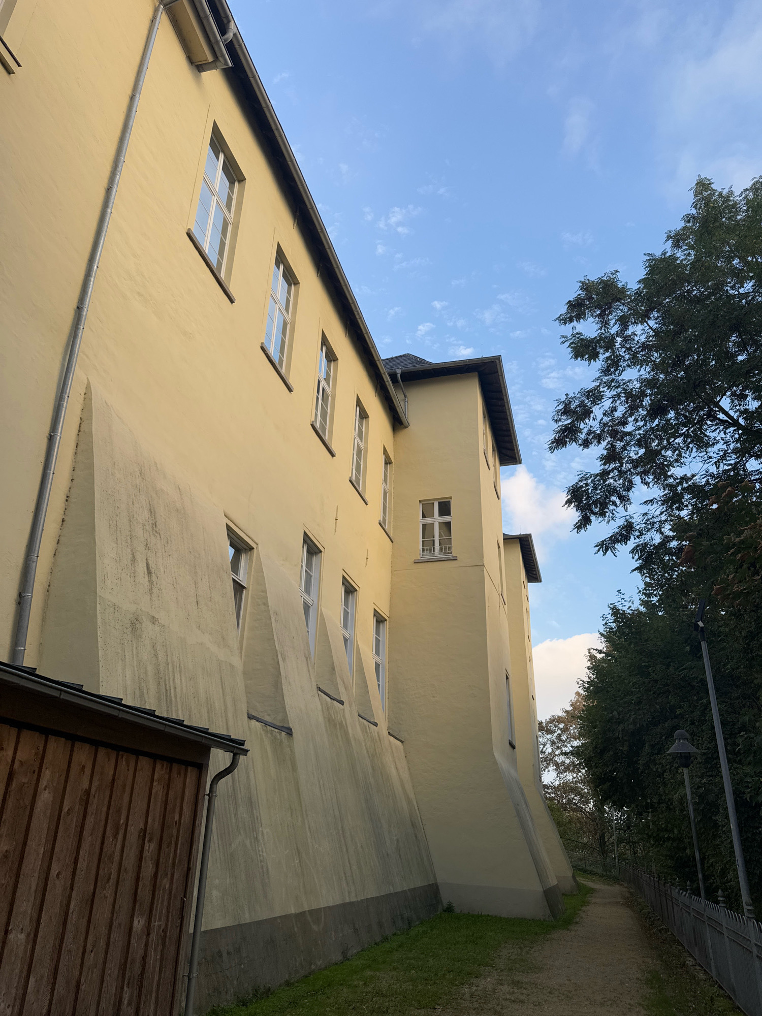 The image shows the exterior of a yellow building with a sloped roof, seen from a low angle. The building has multiple windows with white frames. A drainpipe runs vertically down the side of the building. The wall of the building features several triangular buttresses. In the foreground to the left is a brown wooden structure, possibly a shed or outbuilding, with a dark roof. A gravel path runs alongside the building to the right, bordered by a metal fence and lush green trees. The sky above is blue with scattered white clouds.
This image was taken at the former Franciscan monastery in Cleves, Germany. The building is part of the monastery complex. The perspective suggests the photograph was taken from the pathway leading towards the rear of the monastery buildings.