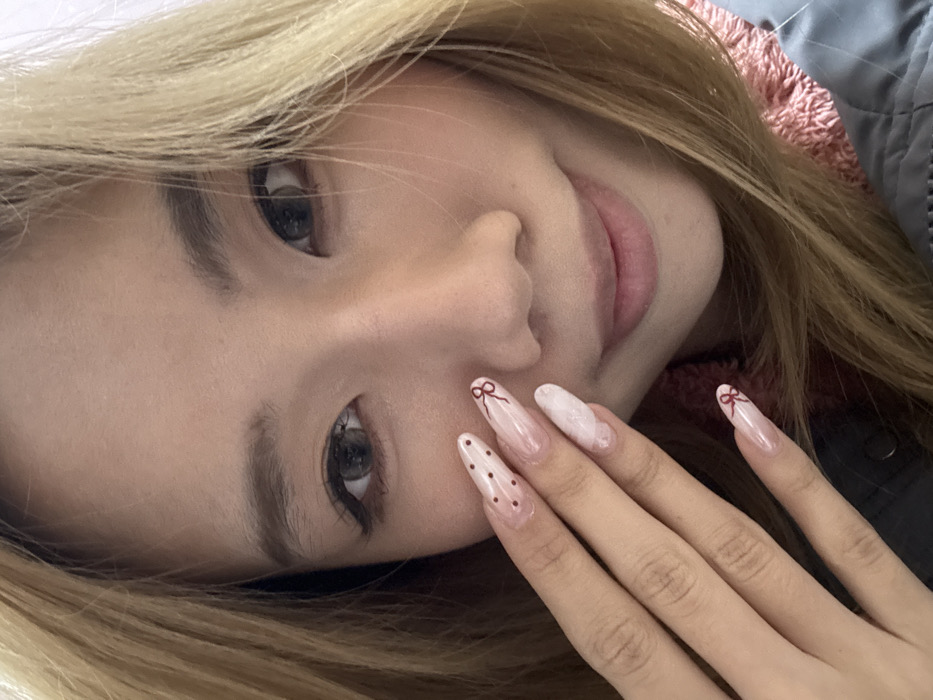This image depicts a close-up portrait of an adult female of East Asian descent, taken in what is identified as Shapingba District, China. The subject's face is oriented directly towards the camera, exhibiting a slight closed-mouth smile. She has long, blonde-toned hair parted on her right side. Her eyes display visible eyeliner and mascara, and her dark irises appear to contain grey-tinted contact lenses. Her left hand is positioned against her left cheek, showcasing five long, almond-shaped artificial fingernails. The index finger nail features dark red polka dots on a clear base. The middle finger nail displays a small dark red bow design on a clear base. The ring finger nail exhibits a milky white, marbled or clouded pattern. The pinky finger nail also has a small dark red bow design on a clear base. The woman is wearing a dark grey outer garment, with a fluffy, reddish-pink inner lining or additional garment visible around her neck and upper chest. The background is uniformly light-colored and out of focus.