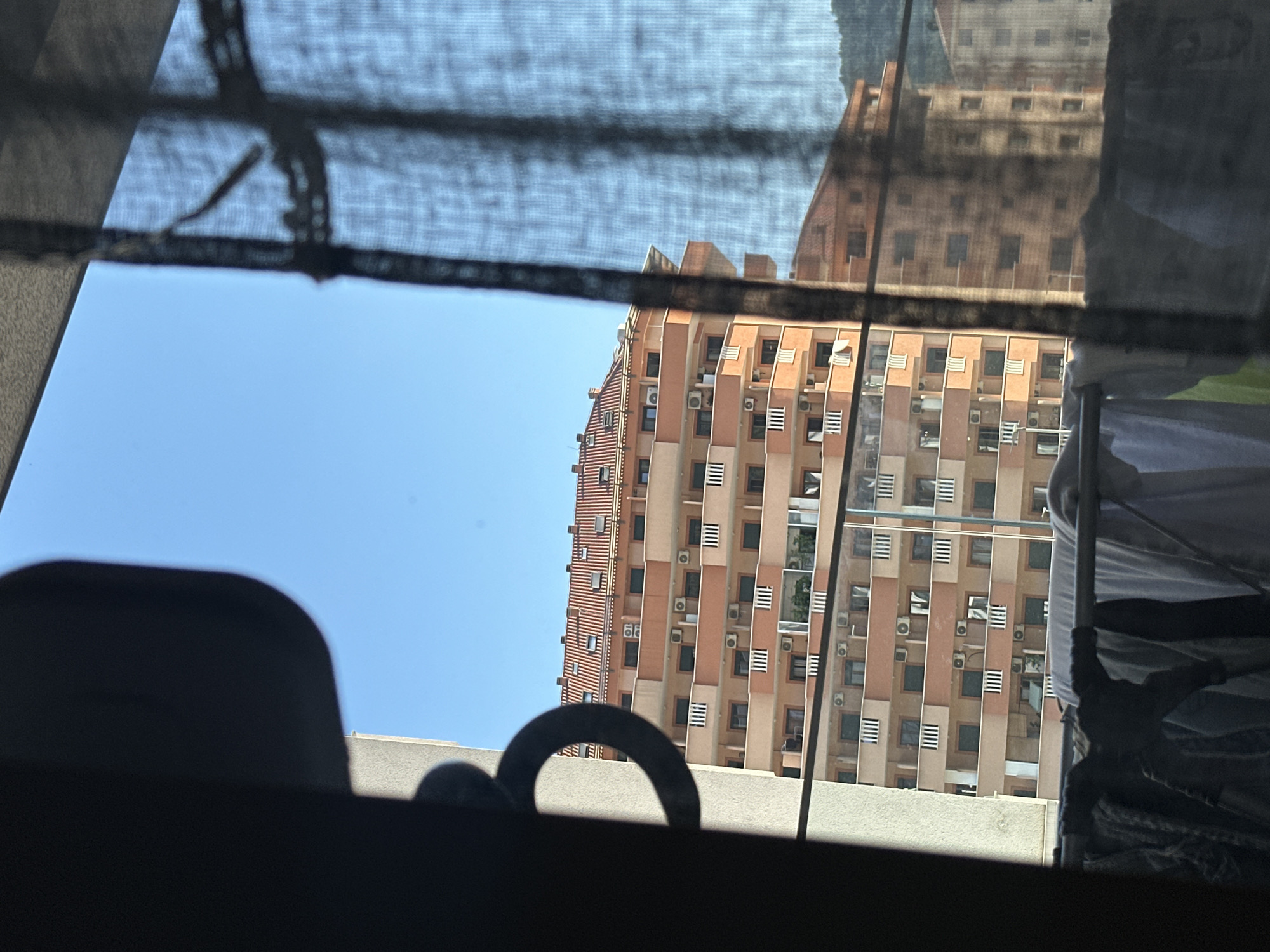 The image depicts a view from a window in Budva, Montenegro. In the foreground, a dark silhouette of a window frame or similar object is visible on the left, with a circular metallic handle attached to it. To the right, a sheer black curtain partially obscures the view. Beyond the curtain, a multi-story apartment building with a terracotta tiled roof is the dominant subject. The building features balconies with white railings and numerous air conditioning units mounted on its exterior walls. The sky is a clear, bright blue, indicating a sunny day. In the lower foreground, clothes are hanging on a clothesline, suggesting an outdoor laundry setup. Another apartment building is partially visible behind the main one. The overall scene is a typical urban residential view, likely from an apartment overlooking other buildings.