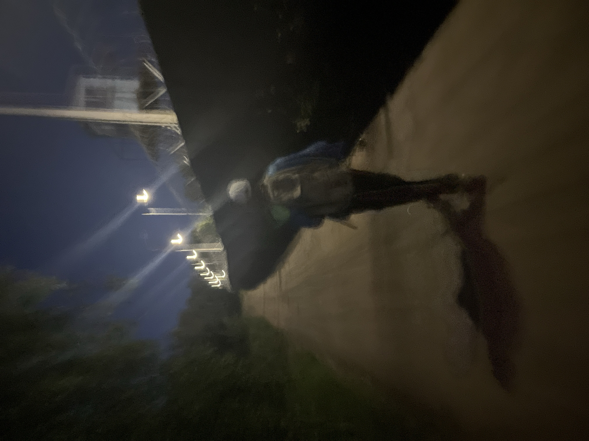 The image shows a person from behind walking down a dirt path at night. The person is wearing a backpack and a blue jacket. There are lights on poles along the right side of the path, and a tall structure with windows is visible in the background, possibly a guard tower. The path is bordered by trees and brush on the left and a dark fence on the right. The sky is dark blue and clear. The scene is consistent with a perimeter fence area, and such areas exist in and around industrial or border regions. Without further context or identifying landmarks, it is not possible to pinpoint the exact location in Chaikovsky, Russia.