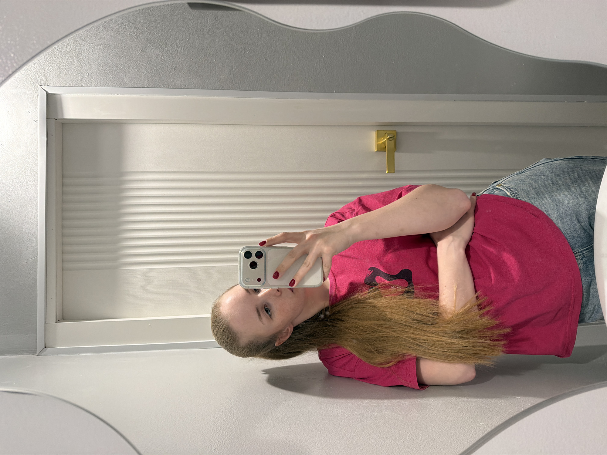 A reflection in a mirror reveals a person with fair skin, auburn hair, and light eyes. They are wearing a pink t-shirt, and blue jeans. They are holding a white smartphone up towards their face. In the background is a white door with horizontal lines and a gold door handle. The walls are grey. The location is a bathroom.