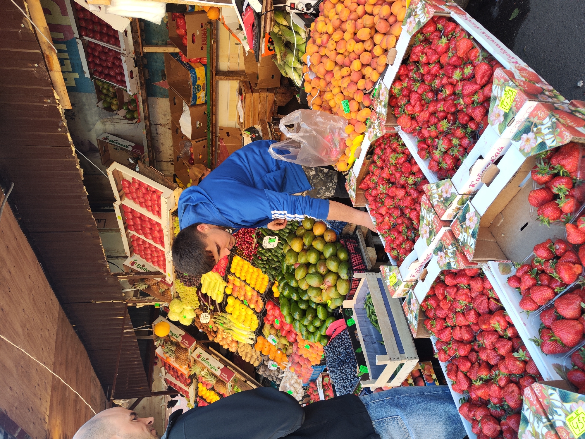 Here is a description of the image.
The image shows a vibrant outdoor fruit and vegetable market. A man in a blue tracksuit is reaching over an array of fresh produce, possibly selecting items for a customer or arranging the display. The market stall is brimming with fruits and vegetables like strawberries, apricots, mangoes, cucumbers, grapes, pineapples, and more. Boxes of strawberries are prominently displayed, and some bear price tags. The market setting suggests an urban environment, and the presence of Russian text may place the location in Saint Petersburg, Russia. Patrons are visible in the foreground and background. The image captures the hustle and bustle of a busy market.