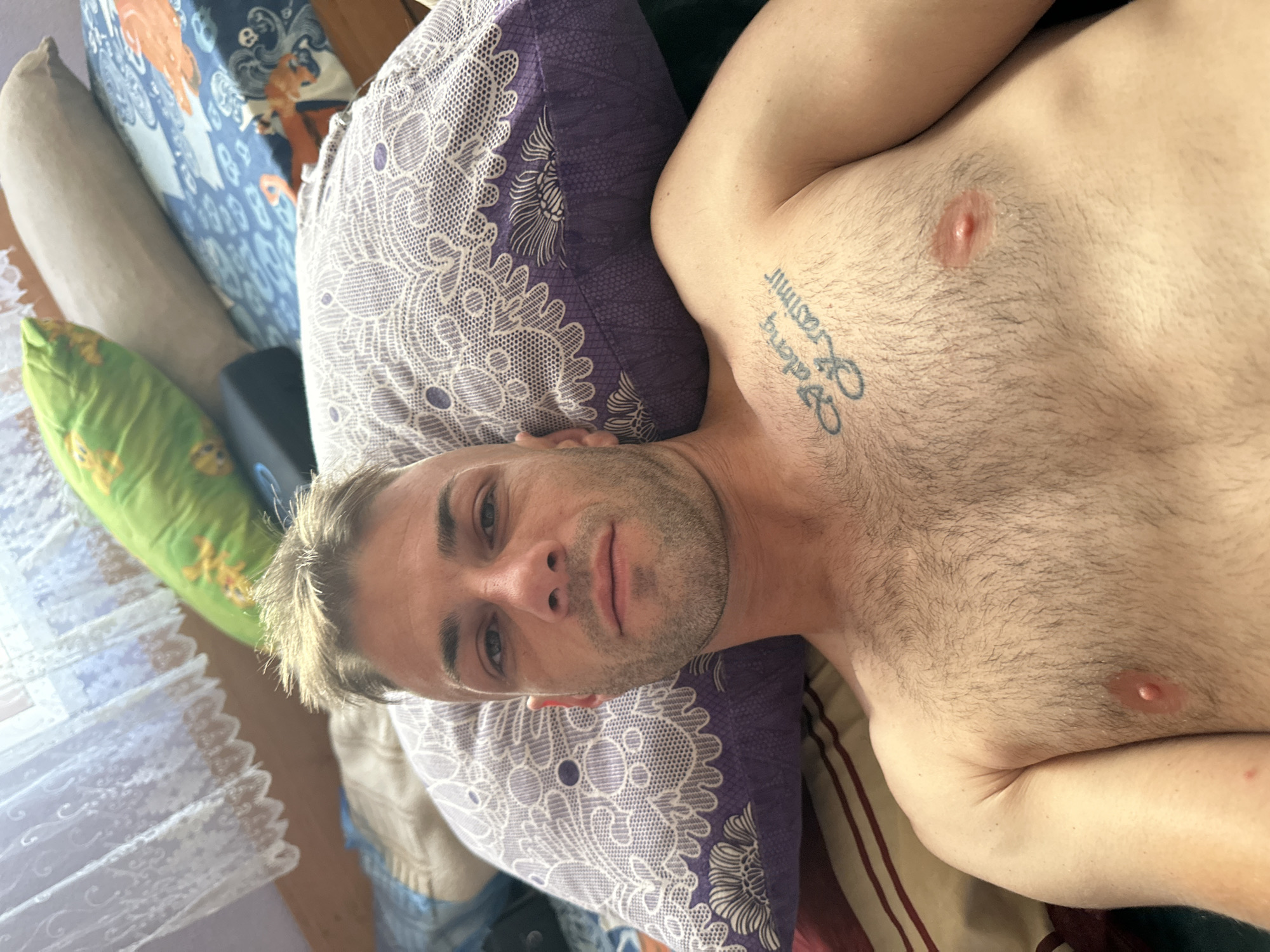 A man is lying on a bed, looking at the camera. He has short, messy blonde hair and a beard. He is shirtless, revealing a tattoo on his chest that reads "Valeri Krasimir". The bed appears to be in a bedroom, with pillows and blankets visible. There are white lace curtains on a window in the background. The scene suggests a casual or private setting. The image does not provide any information about the city of Cherven Breg, Bulgaria.