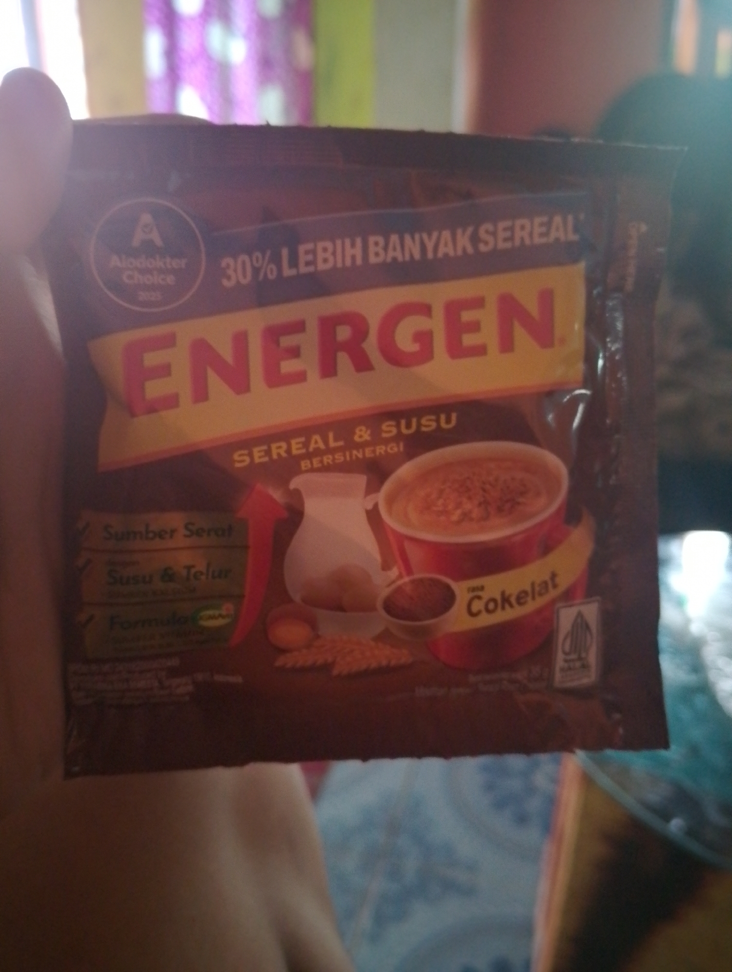 The image shows a hand holding a chocolate-flavored instant cereal and milk drink sachet. The sachet is primarily brown with a yellow and red label featuring the brand name "ENERGEN". Text on the sachet includes "30% LEBIH BANYAK SEREAL" (30% MORE CEREAL), "Sereal & Susu Bersinergi" (Cereal & Milk Synergize), "Sumber Serat" (Source of Fiber), "Susu & Telur" (Milk & Egg), "Formula SMART", and "Cokelat" (Chocolate). There is also a circular logo with an "A" and text "Alodokter Choice 2025". A small halal certification logo is visible on the bottom right. The background is blurred and appears to be indoors, possibly a room with a patterned curtain. There are no clear indications of the specific location in Purwakarta, Indonesia, other than the assumed context.