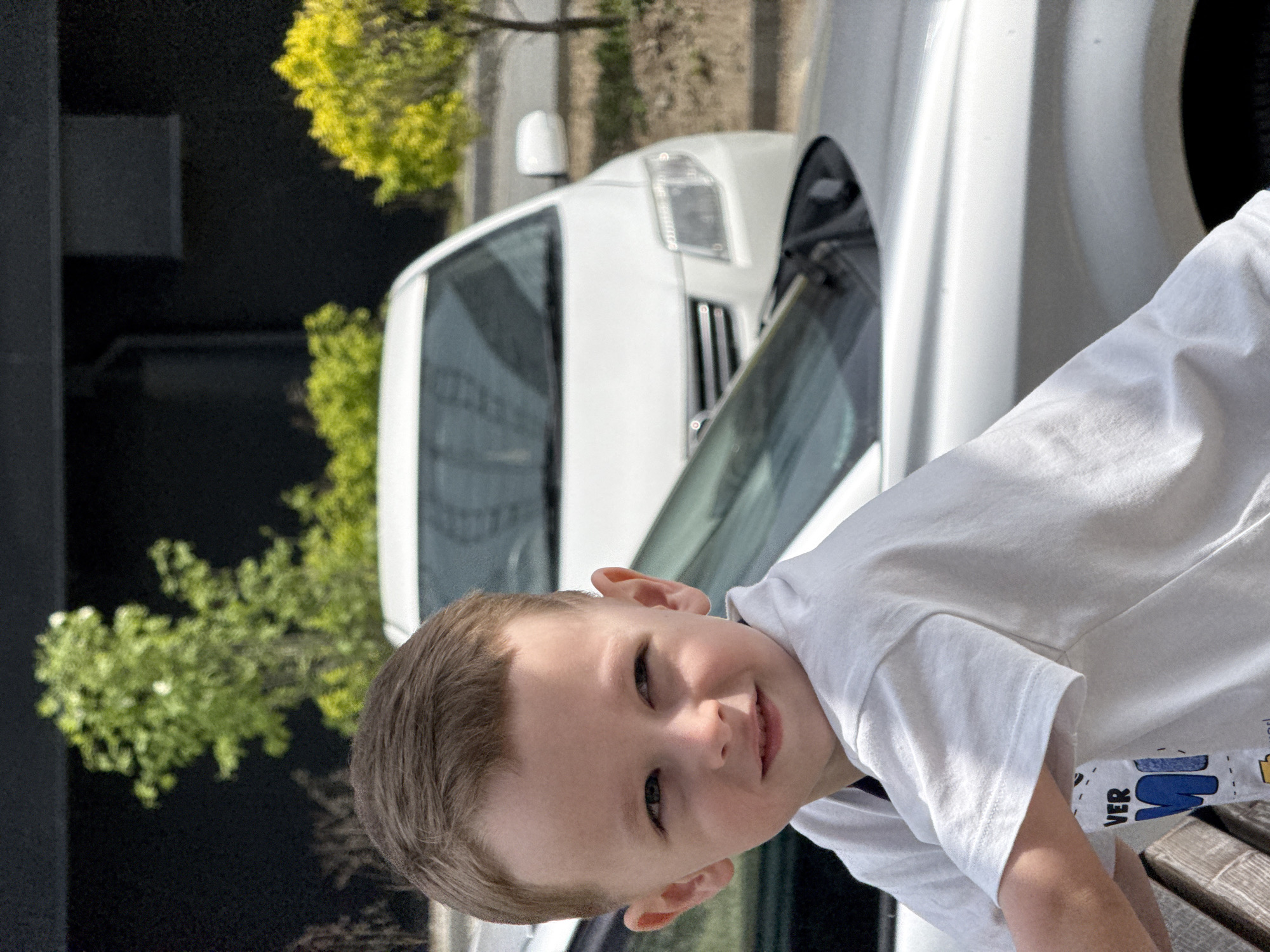 A young boy with short brown hair, wearing a white t-shirt, is sitting on a wooden bench. He is looking at the camera with a slight smile. Behind him, there is a white car parked on a street. In the background, there are trees and a dark building. The image appears to be taken outside, possibly in a city environment, although the specific location in Vladivostok, Russia, cannot be determined with the given information.