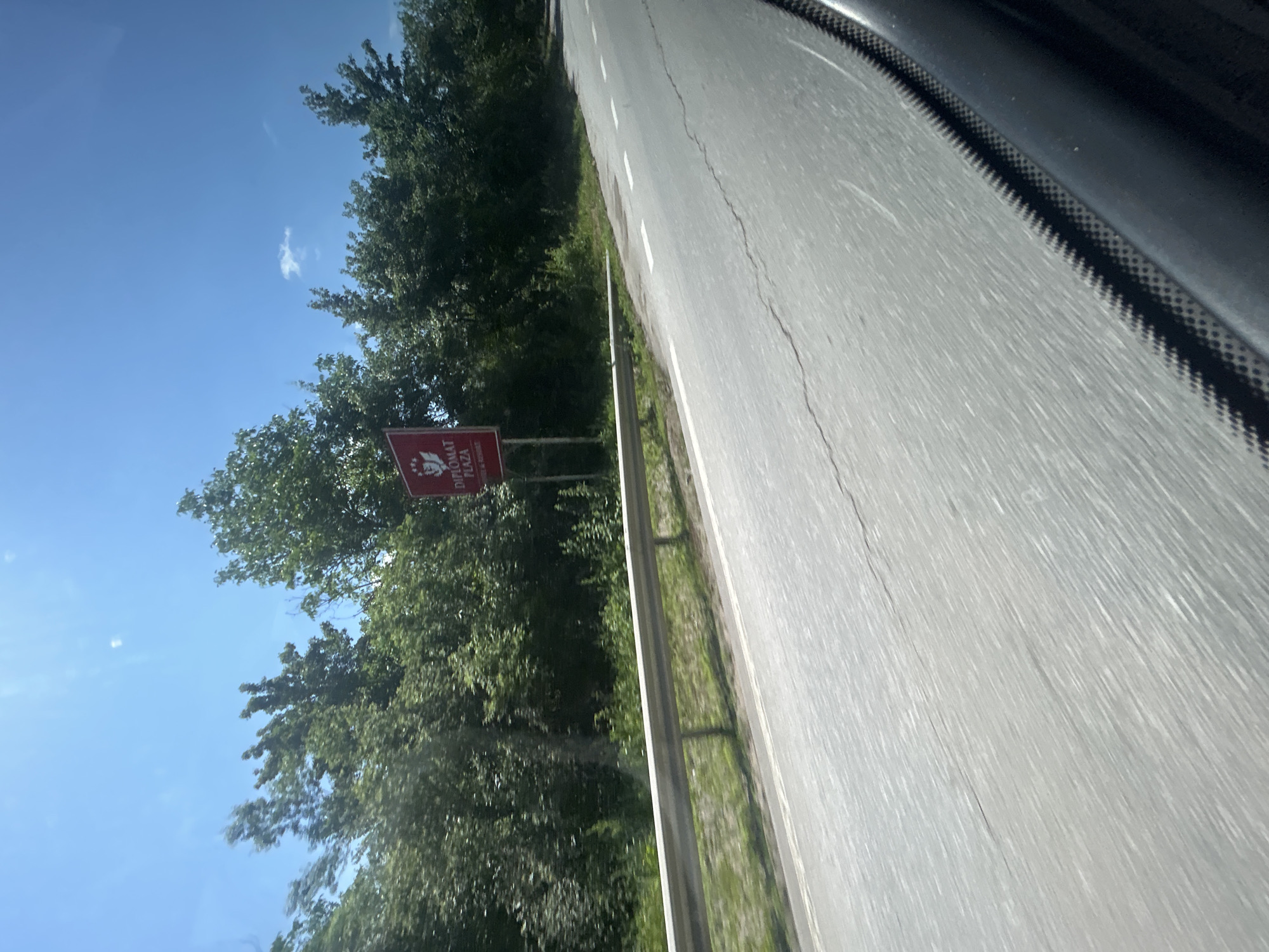 This image captures a bright daylight scene from the perspective of a moving vehicle, looking slightly forward and to the left of the direction of travel. The sky is a clear, vibrant blue with a few scattered white cumulus clouds. Lush green deciduous trees and dense bushes line the left side of the road, forming a natural barrier. In the mid-ground, a red rectangular sign, mounted on two posts, is visible against the foliage. The sign features a white crest-like logo at the top, above the white text "DIPLOMAT PLAZA" in a larger font, followed by "HOTEL & RESORT" in a smaller font. A silver metal guardrail runs parallel to the asphalt road on the left side, separating the road from the vegetated embankment. The two-lane road is paved with grey asphalt, exhibiting visible cracks and white dashed lane markings. In the bottom right corner, a portion of the vehicle's interior is visible, characterized by a dark, textured surface, possibly a dashboard or window frame with a carbon fiber-like pattern, indicating a close-up perspective from within the car. The motion blur on the road and guardrail suggests the vehicle is in motion. The location is Unknown, Unknown.