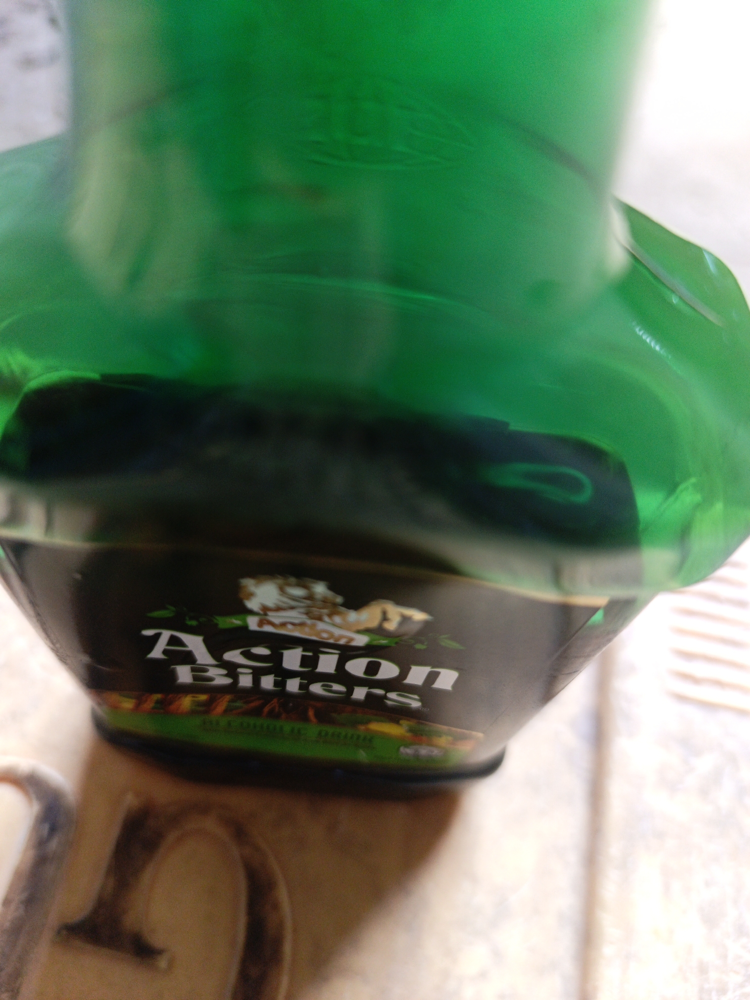 The image shows a partially visible bottle of "Action Bitters" placed on a surface with a tiled pattern. The bottle is green and has a black label with white lettering. The label also includes an image of a lion. The location is Unknown, Unknown.