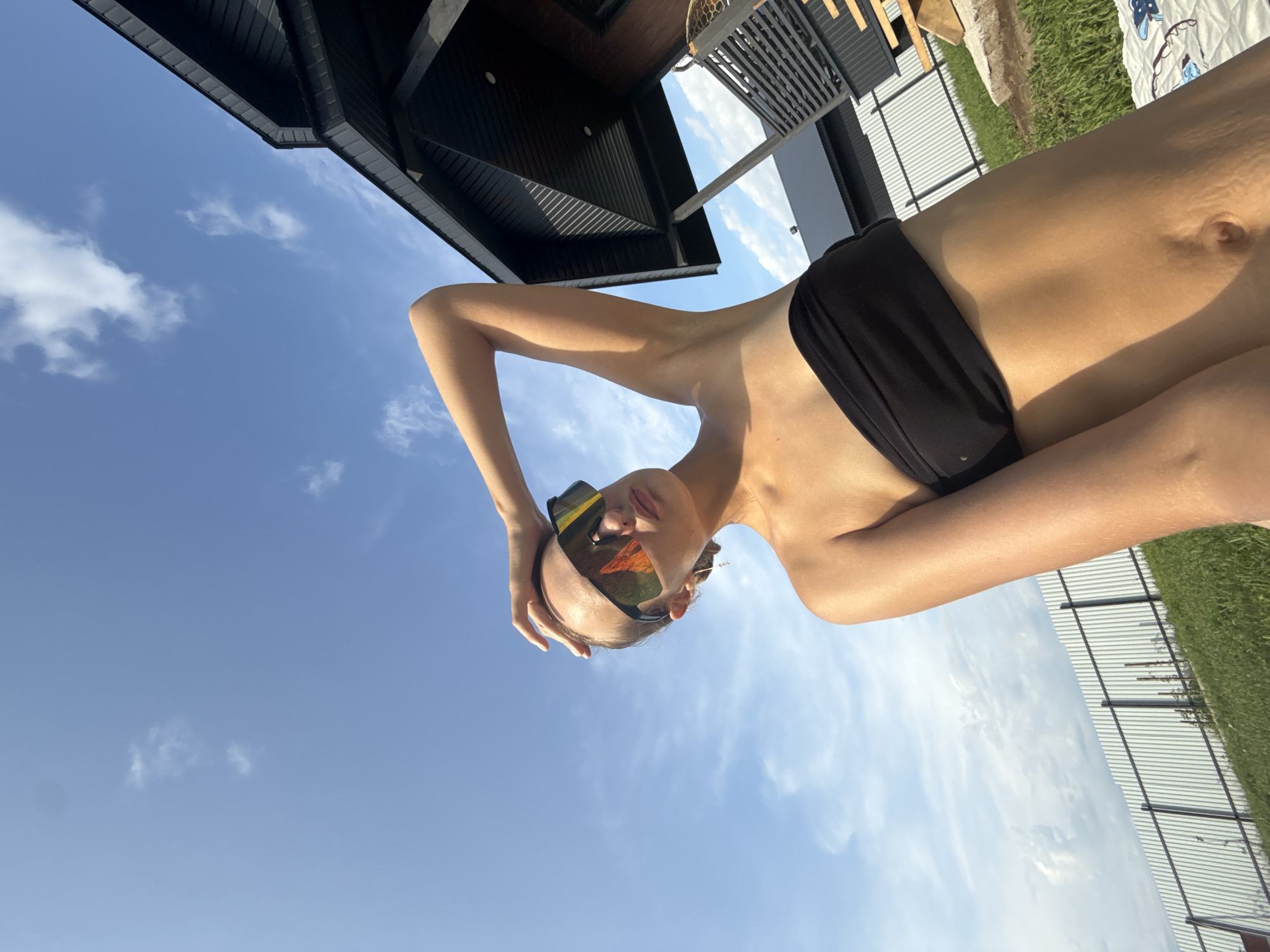 In a low-angle, wide shot, a young woman with blonde hair pulled back is seen from the waist up, outdoors under a bright blue sky with scattered clouds. She is wearing large, reflective sunglasses that obscure her eyes and a black strapless bikini top. Her right arm is raised, with her hand resting on her head. The background features the dark, modern architecture of a house with a steep roofline to the right, and a long, light-colored metal fence running horizontally across the middle ground. Green grass is visible in the foreground. The scene appears to be in a suburban or rural setting, possibly in Podshivalovo, Russia, given the characteristic fence and house style. The lighting suggests a sunny day.
