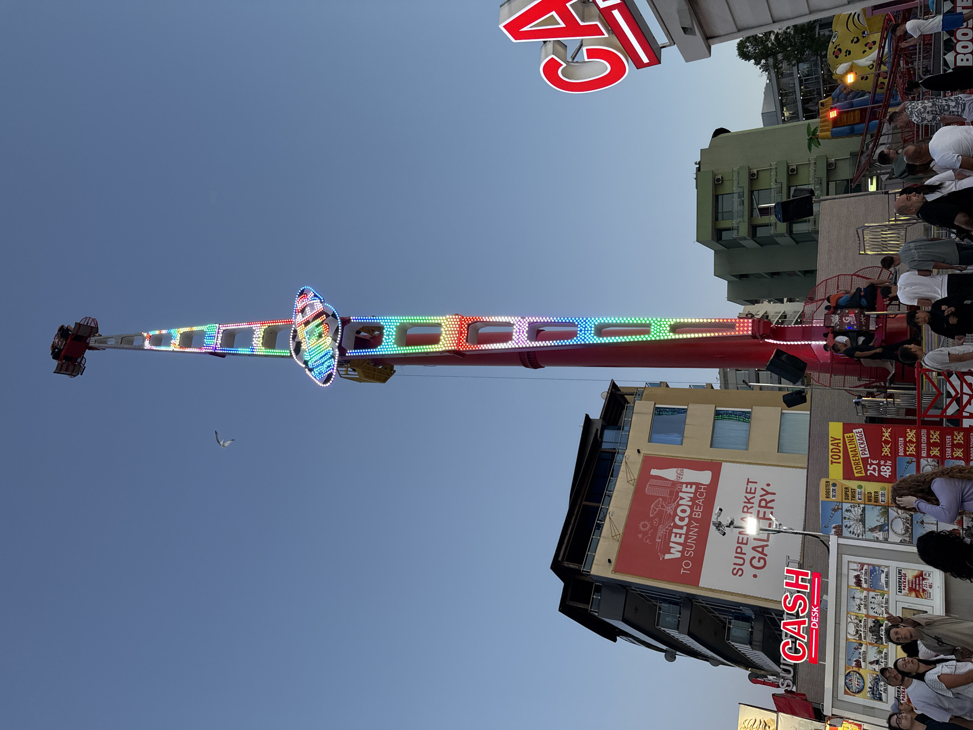 The image depicts an amusement park scene on a clear, pale blue sky, likely in Nesebar, Bulgaria. Dominating the center is a tall, red amusement park ride adorned with colorful LED lights in a rainbow pattern, reaching high into the sky. The ride has multiple levels and appears to be in operation, with people visible on the lower platforms.
In the background, several buildings are visible. To the left, a multi-story building with a large red billboard displaying "WELCOME TO SUNNY BEACH SUPERMARKET GALLERY" stands prominently. To the right of this building, another structure with balconies and windows is present. Further to the right, a vibrant red and white sign reading "CASH" is illuminated. Behind these, several other buildings, including a green-paneled structure, are visible.
In the foreground, a crowd of people is gathered, many looking up at the amusement rides. Some are standing near a ticket booth or information stand with various ride advertisements. The atmosphere suggests a lively, outdoor entertainment venue. A seagull is captured in flight against the sky, adding a natural element to the manufactured environment of the amusement park.