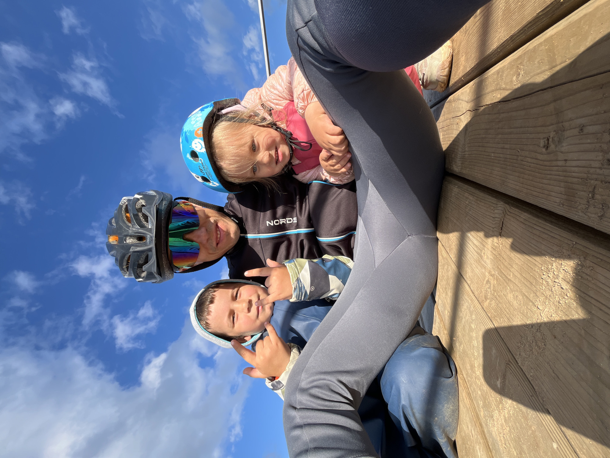 This is a low-angle, wide shot of three people outdoors against a bright blue sky with scattered white clouds. The subjects are positioned in the lower half of the frame, with the upper portion dominated by the sky. They appear to be seated or resting on a wooden surface, possibly a deck or pier, which is visible in the lower right quadrant. The wood is weathered and appears to have some dirt or debris on it.
The subjects are:
1. An adult with a helmet and goggles, appearing to be taking the photo. They are smiling and looking towards the camera.
2. A young child wearing a pink jacket and a blue helmet with colorful cartoon characters on it. They are looking towards the camera with a slight smile.
3. A younger child wearing a blue hooded jacket and making a hand gesture with both hands, appearing to be looking playfully at the camera.
The overall mood is happy and playful, suggesting a family outing. The bright sky indicates it is daytime, likely sunny with fair weather. The clothing suggests a cooler climate, potentially autumn or spring, given the jackets and helmets, which might be for cycling or another outdoor activity.
There is visible text on the adult's black jacket which reads "NORDS" with a blue accent.