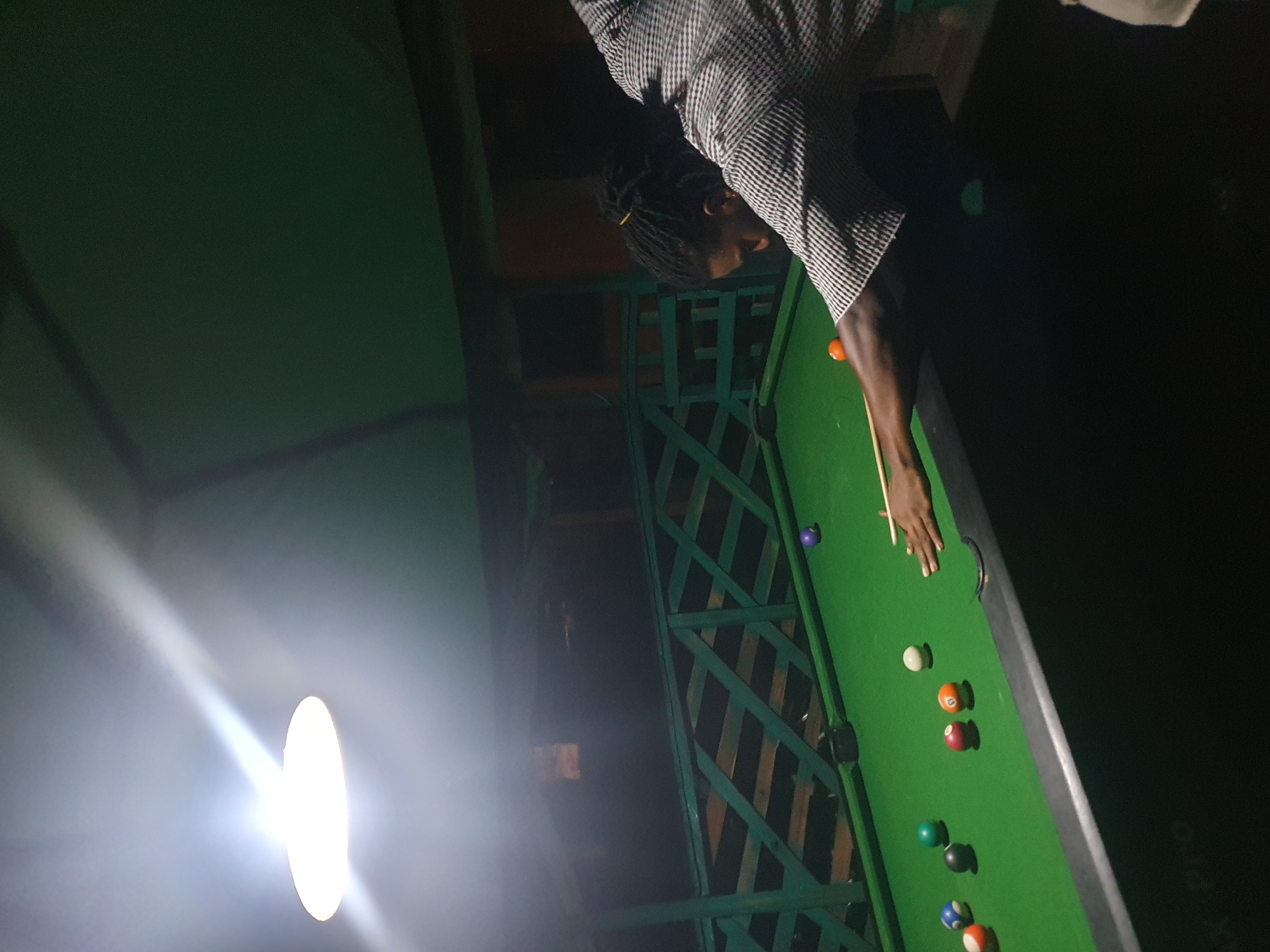 A man with dreadlocks tied back with a yellow band is playing pool. He is wearing a checkered shirt and is in the process of striking a ball with a cue stick. The pool table is green and has several balls scattered across it, including a purple ball, a white ball, and several striped and solid colored balls. The scene appears to be outdoors, possibly a covered patio or open-air bar, with a green wooden railing in the background. A bright overhead light illuminates the table. The image may have been taken in Lokoja, Nigeria, given the context provided.