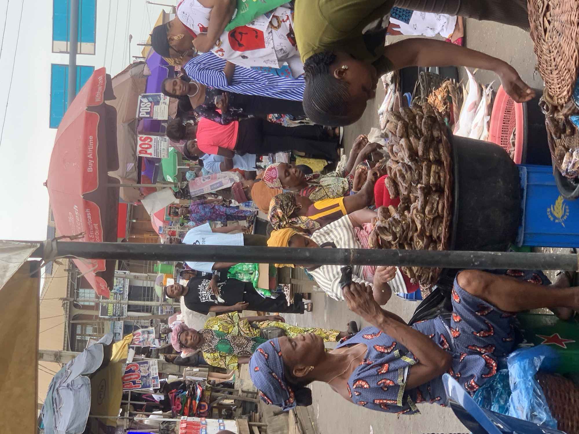 The image shows a bustling marketplace scene, likely in Ikorodu, Nigeria. Numerous people are visible, primarily women, engaged in buying and selling. Several women are seated on the ground behind large baskets overflowing with what appears to be dried fish or smoked seafood. One woman in an olive green shirt is leaning over one of the baskets, inspecting the merchandise. Another woman in a blue and red printed outfit sits nearby, seemingly waiting. Overhead, various red and grey umbrellas are visible, advertising services such as "Buy Airtime" and "Withtaw Tash". POS (Point of Sale) signs are also displayed, indicating money transfer services. The background shows buildings, additional vendors, and other shoppers, suggesting a busy urban market environment. The image conveys a vibrant and active commercial atmosphere.