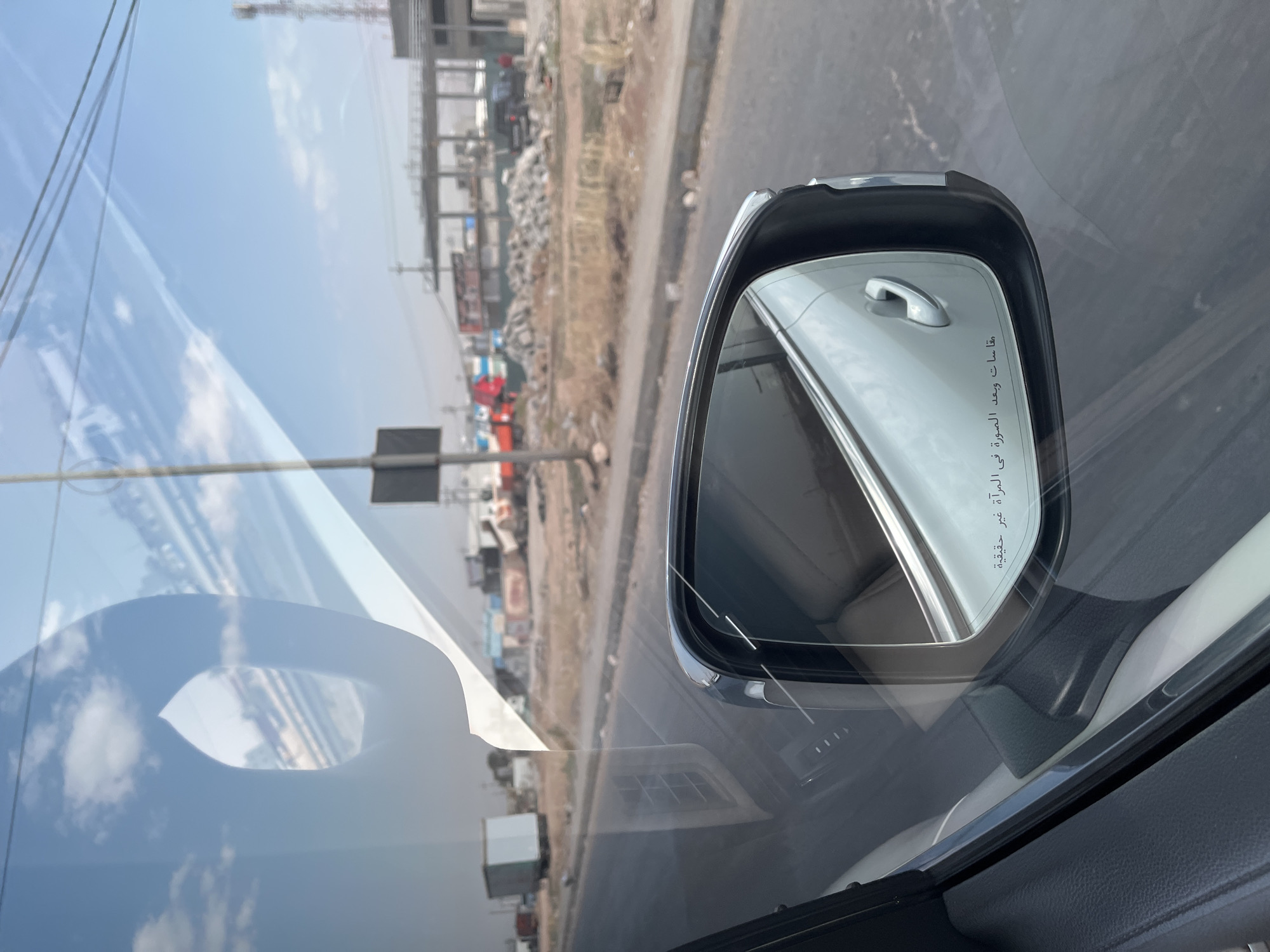 A view from inside a car shows a side-view mirror with a reflection of the car's interior and exterior. On the mirror's surface, Arabic text reads "Objects in mirror are closer than they appear." Outside the car, a road is visible with a sidewalk and a construction site in the background. The construction site features scaffolding, piles of debris, and several vehicles, including trucks and what appears to be an excavator. The sky is blue with scattered white clouds. The overall scene suggests a daytime view of an urban or semi-urban environment undergoing construction.