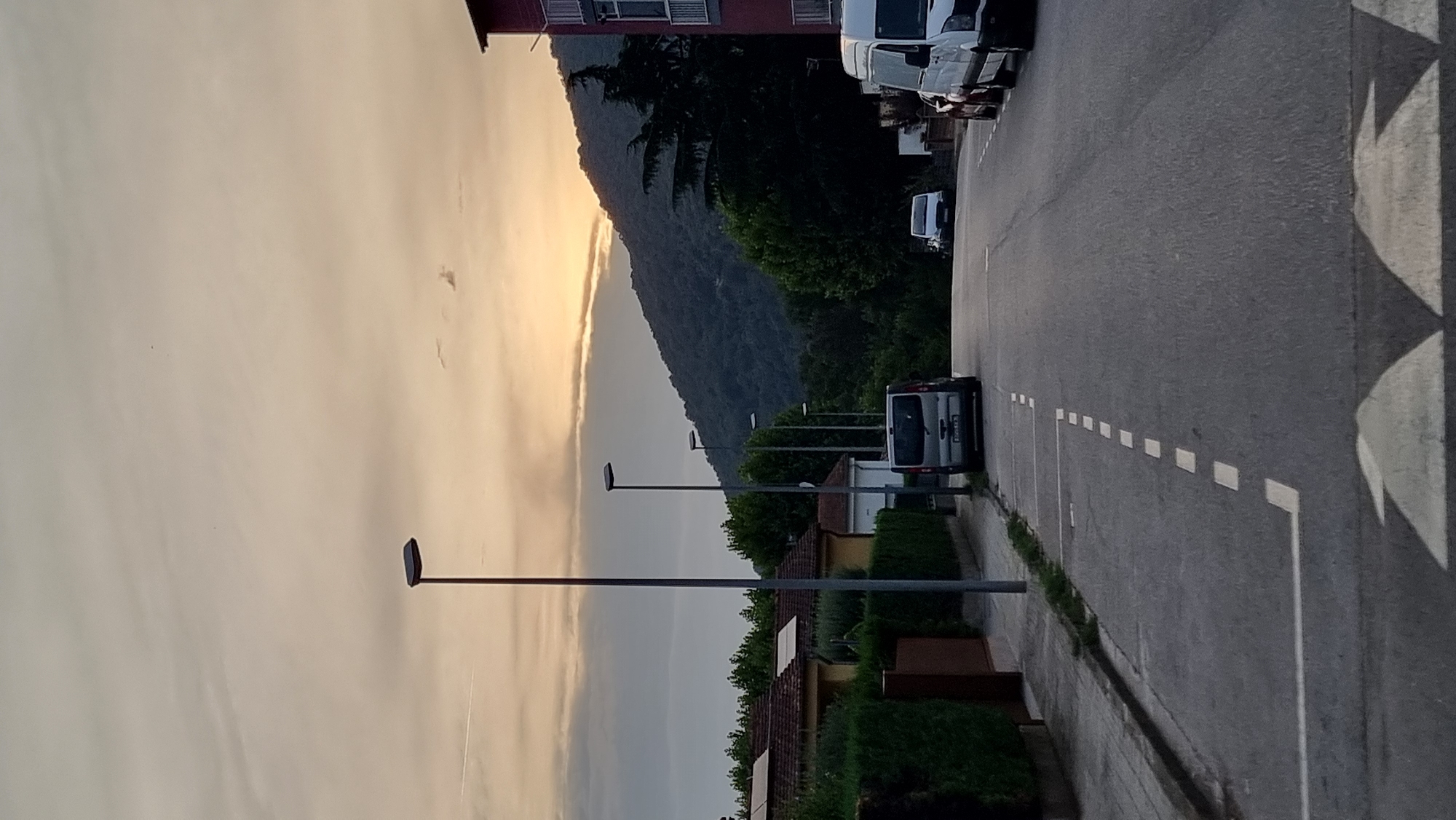 The image depicts a street scene at dusk in Sant Joan les Fonts, Spain, with a mountain range visible in the background. The sky is overcast with soft, muted colors of orange and yellow from the setting sun filtering through the clouds. A street lamp stands on the left side of the road, and several more are visible along the street leading into the distance.
The street itself is paved and has white dashed lines marking parking spaces and solid white lines indicating the edge of the road. Several vehicles are parked or driving on the street. On the left, a silver minivan is parked. Further down the street, a white van is visible. On the right side of the road, a white van and a dark-colored car are parked.
The left side of the street is lined with houses and dense green hedges. The houses have red tile roofs, and some appear to have solar panels installed. Trees and more dense vegetation are visible behind the houses and along the base of the mountain. The mountain itself is covered in dark green trees and appears to be a significant feature of the landscape. A building with a red facade and balconies is visible on the far right side of the image. The overall impression is a quiet, residential area during the evening hours.