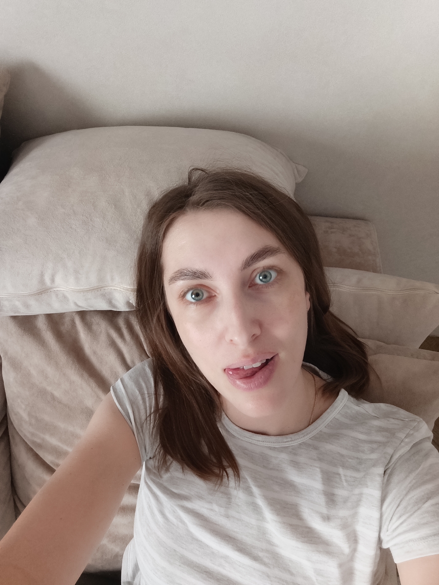 A woman with brown hair and blue eyes is lying on a bed. She is wearing a grey and white striped t-shirt. She is sticking her tongue out. There is a pillow behind her head. The location is unknown, but based on the query, it is possibly Chaikovsky, Russia.