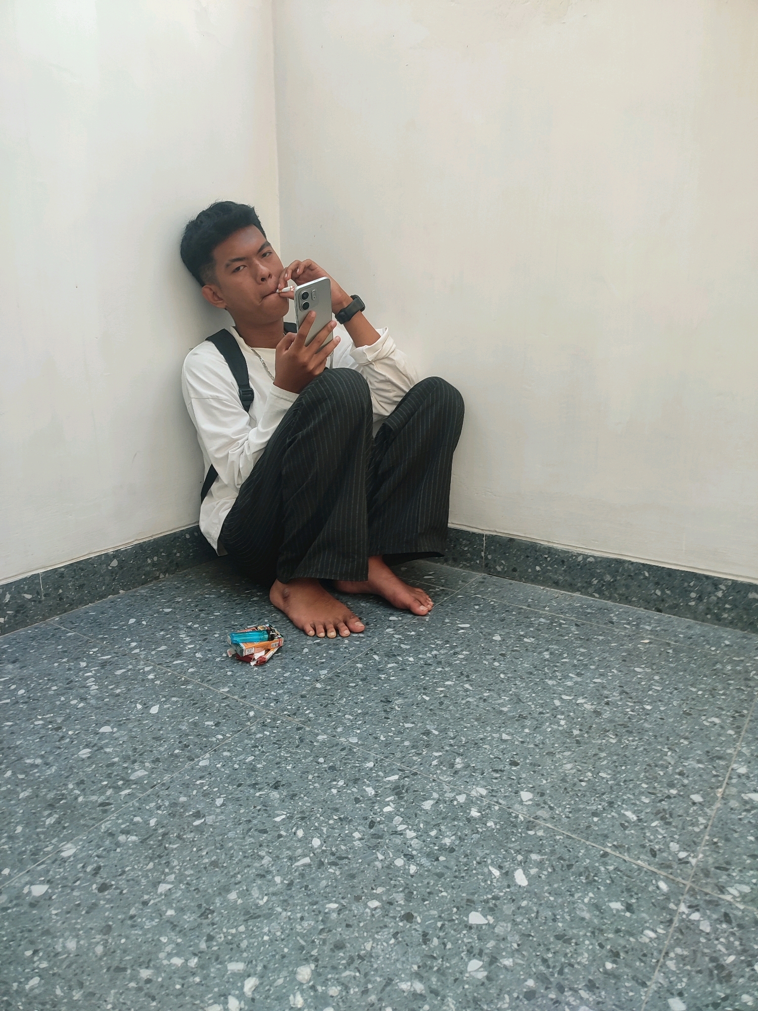 A young male with dark hair is sitting in the corner of a room, leaning against a white wall. He is wearing a white long-sleeved shirt and dark, pinstriped pants. He is holding a smartphone in his right hand and has a cigarette in his mouth. A backpack is visible over his left shoulder. On the floor in front of him are a pack of cigarettes and what appear to be loose cigarette butts. The floor is made of grey terrazzo tiles. The environment suggests an indoor setting, possibly a public space or a room within a building in Trenggalek, Indonesia.