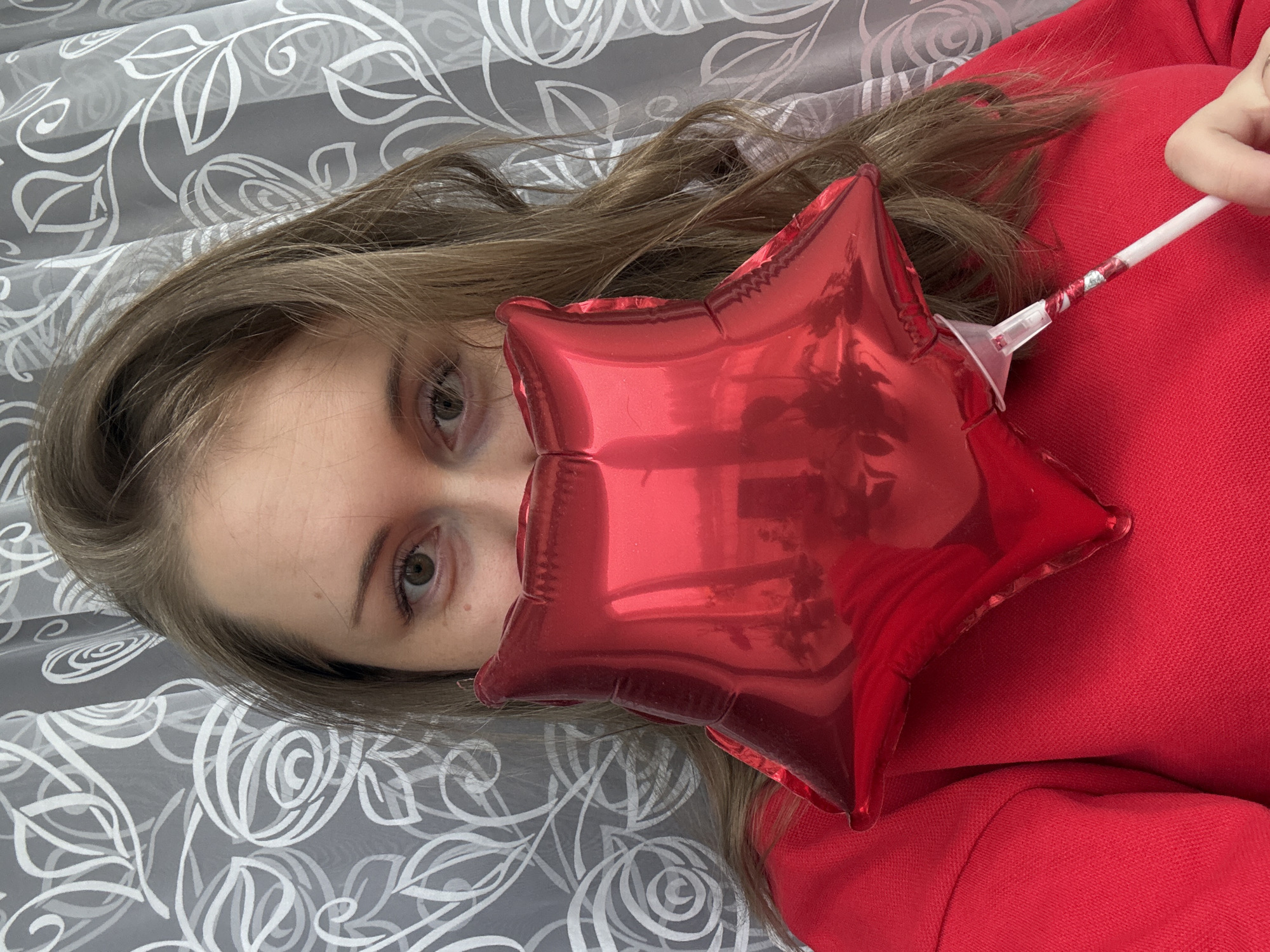 The image, rotated 90 degrees clockwise, presents a close-up view of a person, likely female, partially hidden behind a red star-shaped balloon. Her long, light brown hair frames her face, with only her eyes and forehead clearly visible. Her eyes, appearing to be hazel or greenish-brown, gaze directly at the viewer.
A small, reflective red foil balloon, shaped like a five-pointed star, is positioned in front of her lower face. The balloon is attached to a white stick, which has a decorative red and silver pattern near the balloon's base. A hand, presumably hers, is visible in the upper right, holding the balloon stick.
The person is wearing red clothing, which appears to be a textured fabric, possibly a sweatshirt or knit top, seen on the right side of the frame. The background features a grey fabric or curtain adorned with a distinctive white floral or leafy pattern. The setting suggests an indoor environment. There is no visible text in the image. The overall impression is one of a playful or casual portrait.