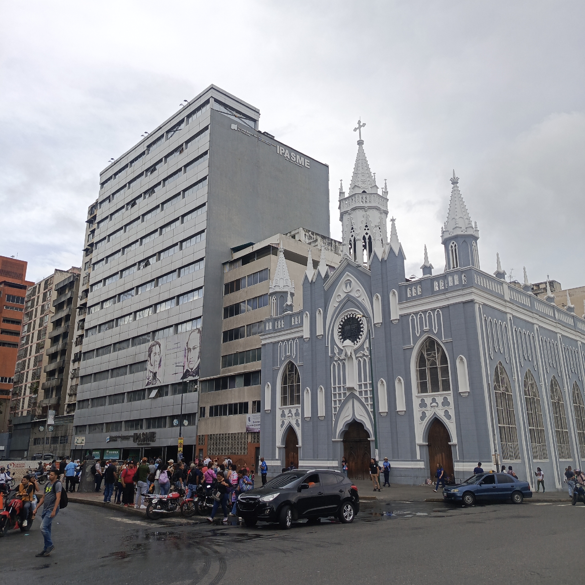 This image depicts a street scene in Caracas, Venezuela, with a prominent grey, multi-story office building on the left and a light blue church with white Gothic architectural details on the right. The sky is overcast. The office building has numerous windows and features a large banner with the text "IPASME" and portraits of two men. In front of the office building, there is a sidewalk with a crowd of people, many of whom are gathered near the entrance of what appears to be a pharmacy or store, indicated by signage for "FARMA." There are also several motorcycles parked and people interacting on the street. A black SUV is parked in the street in front of the church. To the right of the SUV, a blue sedan is also parked. Pedestrians are walking on the sidewalk and crossing the street. The street appears wet in places, suggesting recent rain. The overall impression is a busy urban environment with a mix of commercial and religious architecture.