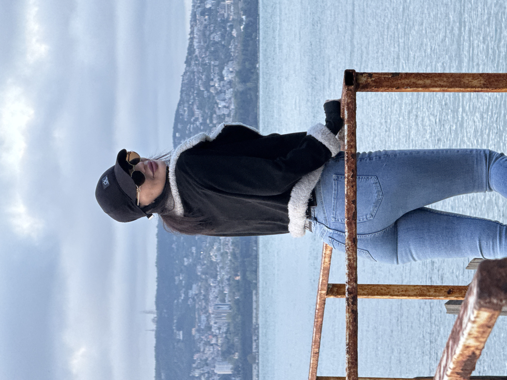 A woman stands on a rusted metal structure, potentially a staircase or platform, near a body of water. She wears a black jacket with a white fur-like trim, blue jeans, a black baseball cap with a logo, and sunglasses. The environment behind her appears to be a coastal city, possibly Varna, Bulgaria, with buildings visible on the hillsides. The sky is overcast. The woman leans against the metal structure, looking towards the sky. The focus is on the woman and her immediate surroundings.