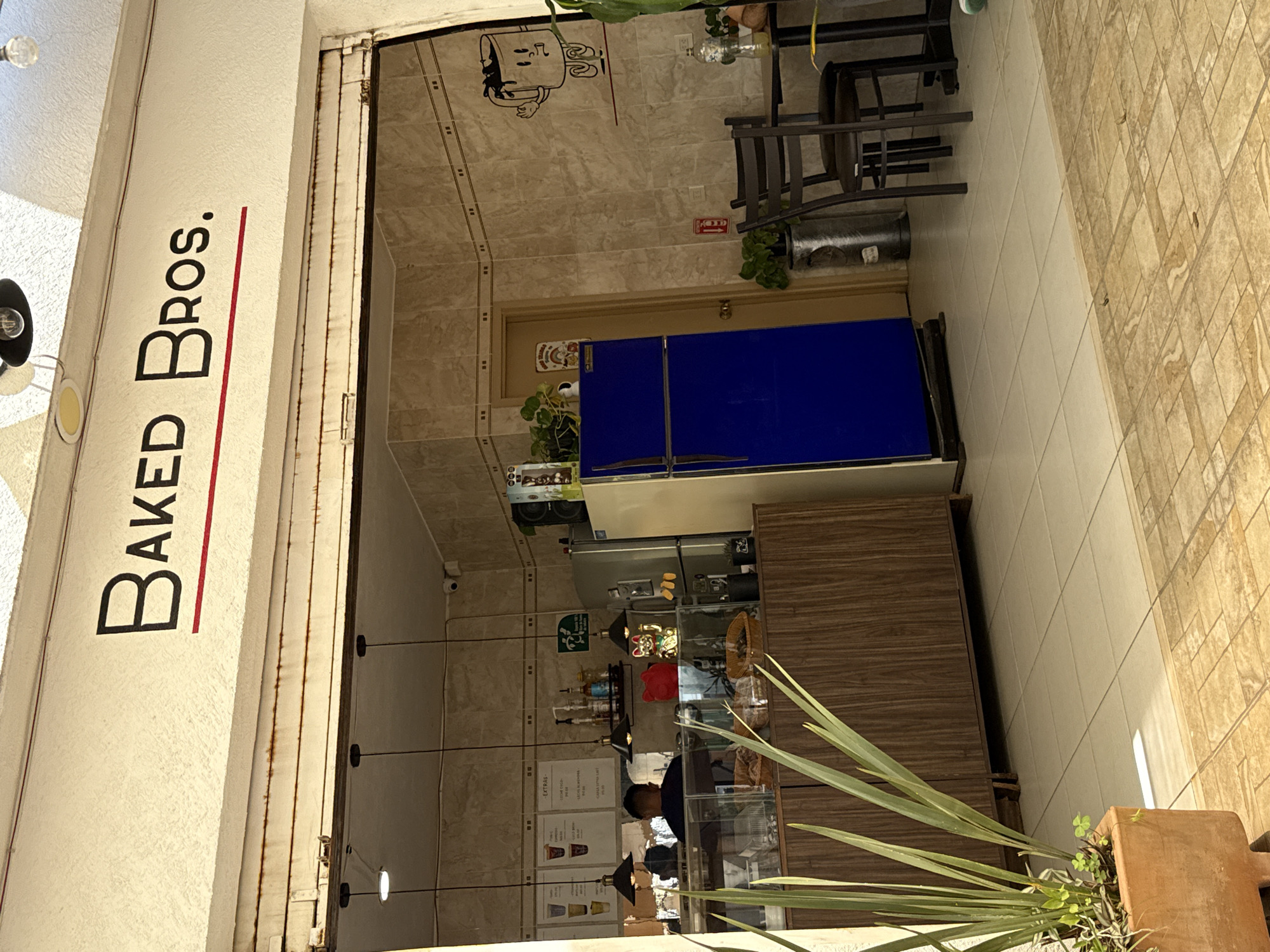 This image depicts the open storefront of a business named "BAKED Bros." located in San Bernardino Tlaxcalancingo, Mexico. The establishment's name is displayed in black capital letters with a red underline on a textured white facade above the entrance. A partially rolled-up, rusty corrugated metal security shutter is visible directly above the open doorway.
The foreground shows an outdoor tiled area with light brown and beige geometric patterns. To the left, a terracotta planter contains tall green leafy plants. The interior floor consists of light beige square tiles.
Inside, a wooden service counter with a glass display case runs across the width of the space, holding various baked goods and a basket. A person, observed from behind, with dark hair and wearing a dark shirt, stands behind this counter. Above the counter, several black pendant lights are suspended from the ceiling.
The interior walls are covered in light beige square tiles. On the left wall, menu boards displaying drinks are visible. Various objects are arranged on and behind the counter, including bottles of syrups, a red stylized cat figurine, and a gold Maneki-neko (waving cat) figurine. A large, two-door bright blue refrigerator stands on the right side of the interior, next to a light brown wooden door. A stainless steel refrigerator unit is positioned to its left. On the tiled wall to the right of the blue refrigerator, a cartoon drawing of a coffee cup with limbs is affixed. A red fire extinguisher is mounted on the wall below this drawing. A small square black table with two dark-colored chairs is positioned on the right side of the interior, with a plastic bottle containing a yellow liquid on the table surface. A silver trash receptacle is visible near the table. A security camera is mounted in the upper right corner of the interior space.