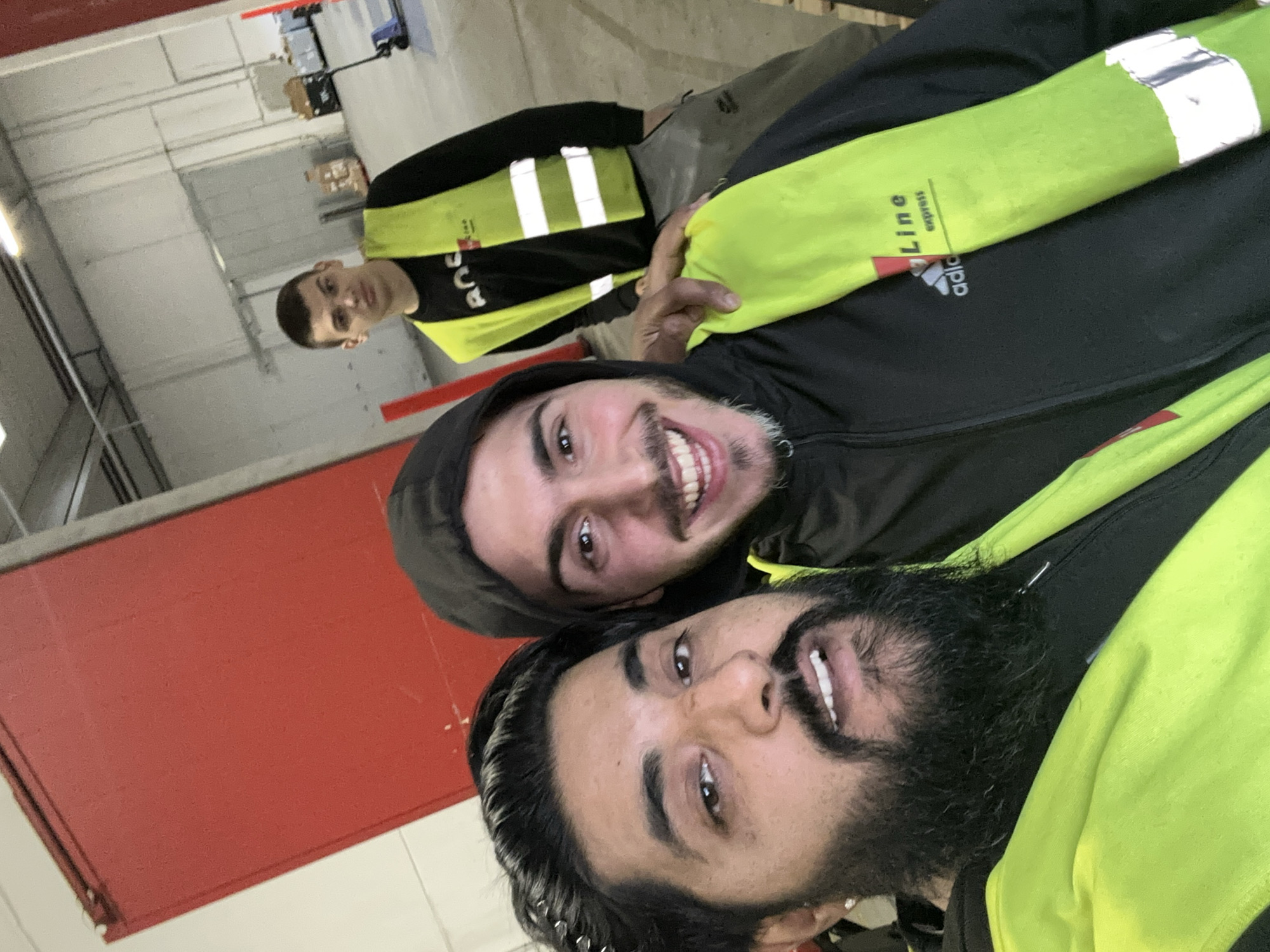 The image shows three men in what appears to be a warehouse or industrial setting. In the foreground, two men are taking a selfie. The man on the left has a beard and is wearing a bright yellow safety vest over a black jacket. The man on the right is also wearing a black jacket, partially covered by a yellow safety vest, and is smiling broadly. In the background, a third man is visible, wearing a yellow safety vest over a black shirt. He is looking towards the camera. The environment is utilitarian, with concrete floors, large red doors, and industrial shelving visible in the background. There are also some pallets and a pallet jack. The overall impression is of a casual moment captured during a workday.