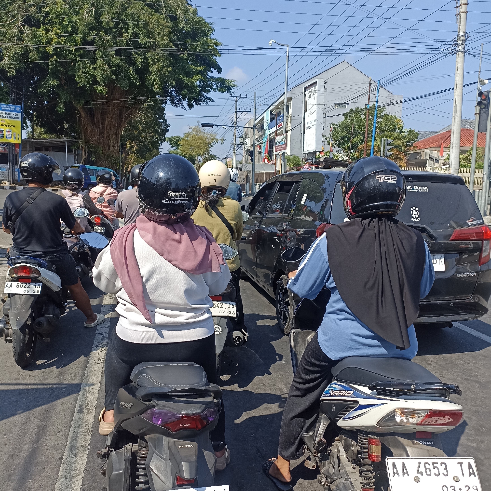 This is a daytime, eye-level, medium shot of a traffic jam on a street in Magelang, Indonesia. The foreground is dominated by the rear of three motorcycles and one car. On the leftmost motorcycle, a person wearing a black helmet and a black t-shirt is visible from the shoulders up. To their right, a person in a white top and pink headscarf, wearing a black helmet and black pants, is seated on a motorcycle with license plate AA 6022 TA. Further right, a person wearing a light tan helmet and a green jacket is on a motorcycle. Behind them, a black car is stopped in traffic. On the right side of the image, a person wearing a black helmet and a blue shirt is on a motorcycle with license plate AA 4653 TA. The background shows buildings, trees, power lines, and a traffic light. The street appears to be paved with asphalt and has white lane markings. The overall atmosphere is that of a typical urban traffic scene under a clear blue sky.