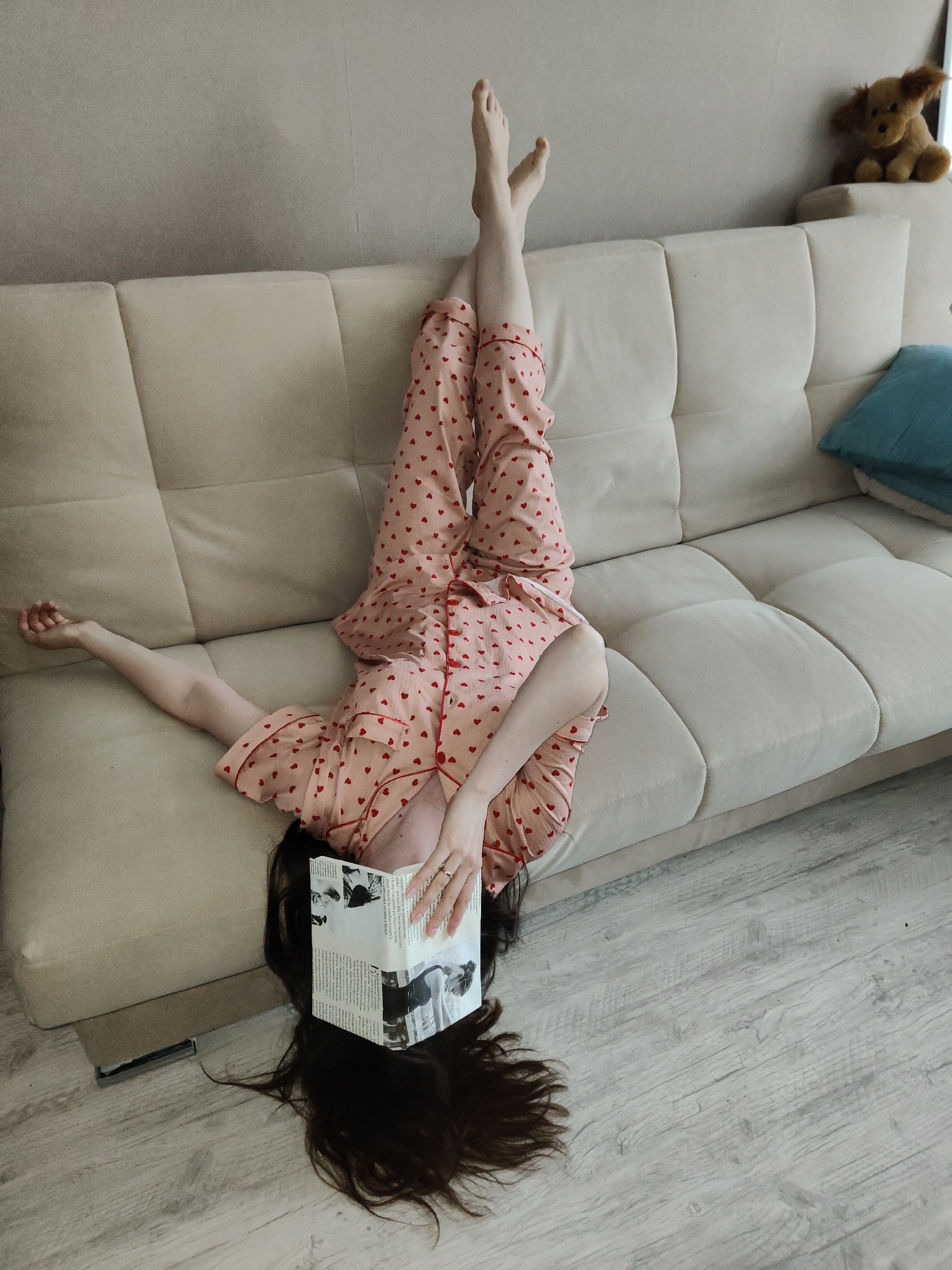 A person is seen lying upside down on a light-colored, tufted sofa indoors. Their head hangs towards the light wood laminate floor, with long dark hair spread out. They are wearing pink pajamas with small red heart patterns. Their arms are relaxed, with one arm extended along the sofa cushion and the other holding an open book over their face, obscuring it. Their legs are extended upwards, bent at the knees, with their bare feet resting against the backrest of the sofa.
The setting appears to be a cozy living space or bedroom. To the right of the person's feet, a brown stuffed animal, resembling a small dog or bear, sits on the back of the sofa. A blue throw pillow is also visible on the far right of the sofa. The wall behind the sofa is plain and light-colored.
The activity is one of casual relaxation, possibly reading, given the book held over the face. The inverted pose suggests comfort and perhaps a playful or whimsical mood. The book contains visible text and black and white images; one discernible word on the right page appears to be "l'histoire," suggesting a non-English language, possibly French. A small, dark rectangular object, possibly a phone or remote, is on the floor near the sofa. The overall atmosphere is serene and informal.