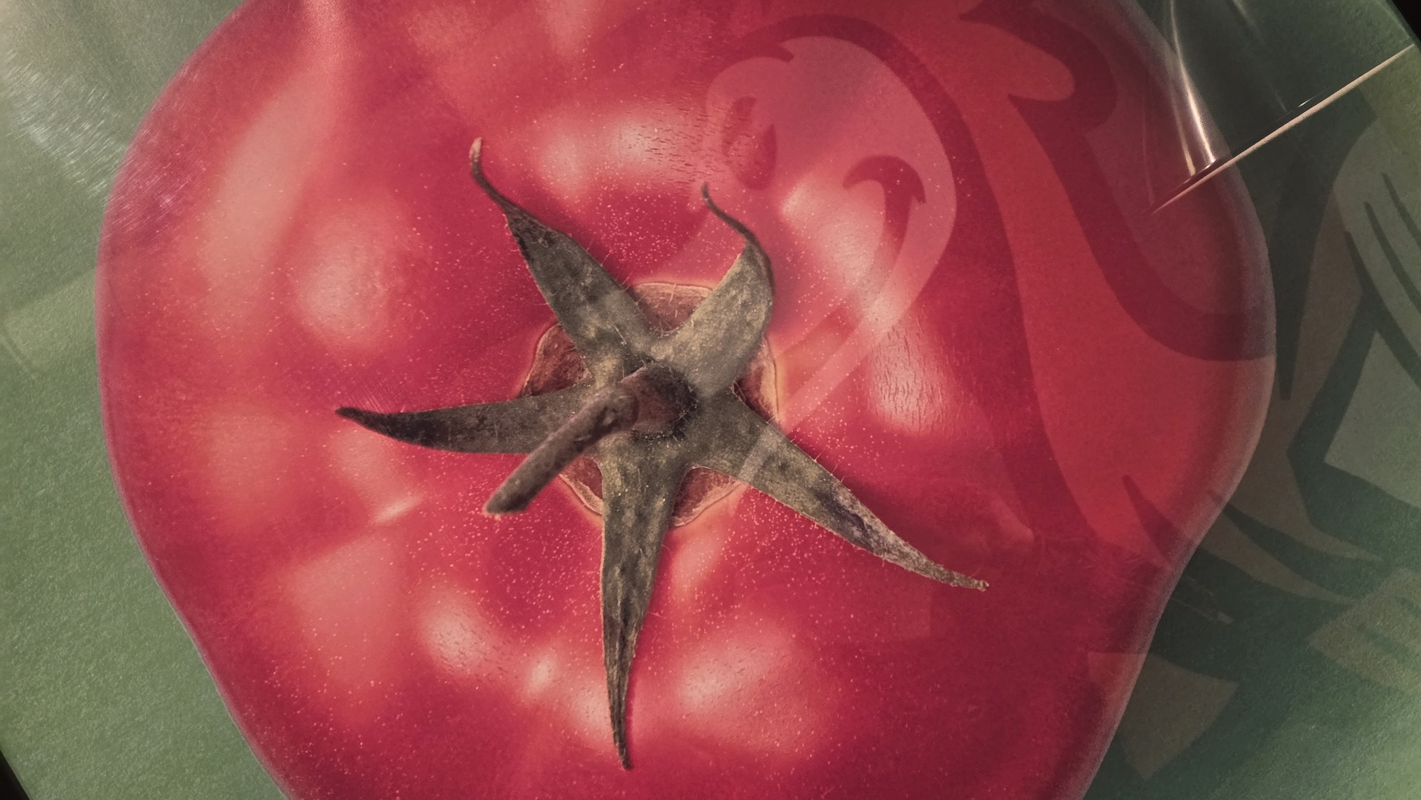The image shows a close-up, top-down view of a red tomato with a green stem. The tomato has a shiny, smooth surface with some light reflections and what appear to be tiny speckles of dust. A thin, white object, possibly a needle or skewer, is inserted into the right side of the tomato. The background is a green surface with a swirling, abstract pattern in a darker shade of green. There are no people or any visible elements that would help in determining its location in Chaikovsky, Russia.