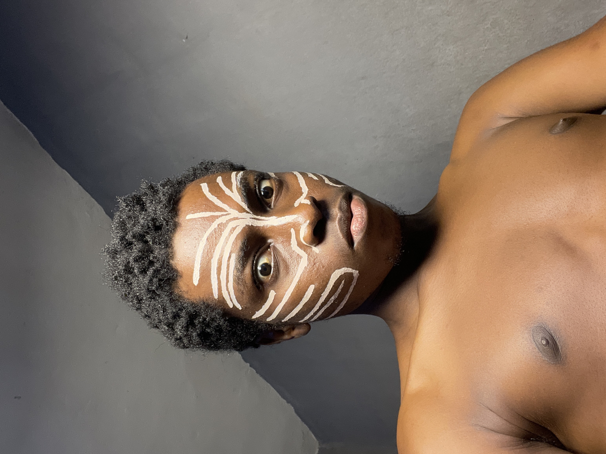 A young Black male with dark, short, textured hair and brown skin is the subject of this chest-up portrait. He has white face paint applied in tribal-like markings across his forehead, cheeks, and nose. His eyes are wide and looking directly at the camera with a serious expression, and his lips are slightly pursed. He is shirtless, showing his chest and shoulders. The background is a plain, gray wall with some subtle shadow work that creates diagonal lines. The setting appears to be indoors. The individual is located in Nkpor, Nigeria.