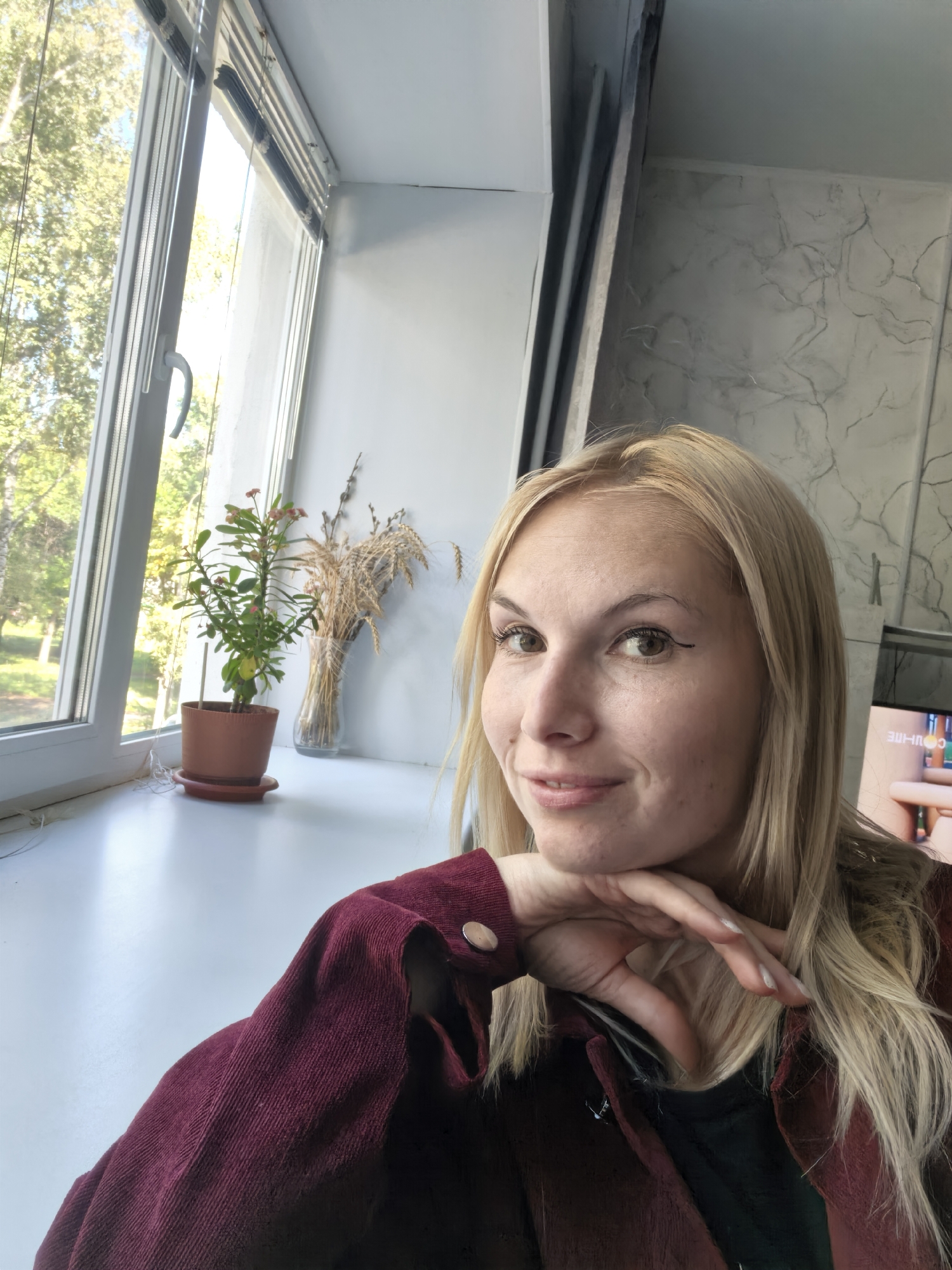 A blonde woman with light brown eyes and subtle makeup, including winged eyeliner, smiles at the camera while resting her chin on her hand. She is wearing a dark red corduroy jacket with silver buttons. The setting appears to be indoors near a window. On the windowsill, there is a small potted plant and a glass vase containing dried wheat and pussy willow branches. Outside the window, trees and foliage are visible, suggesting a possibly temperate climate. The wall behind the woman features a textured gray pattern resembling marble. The image has been taken from a slightly elevated angle. The location is not specified in the image.