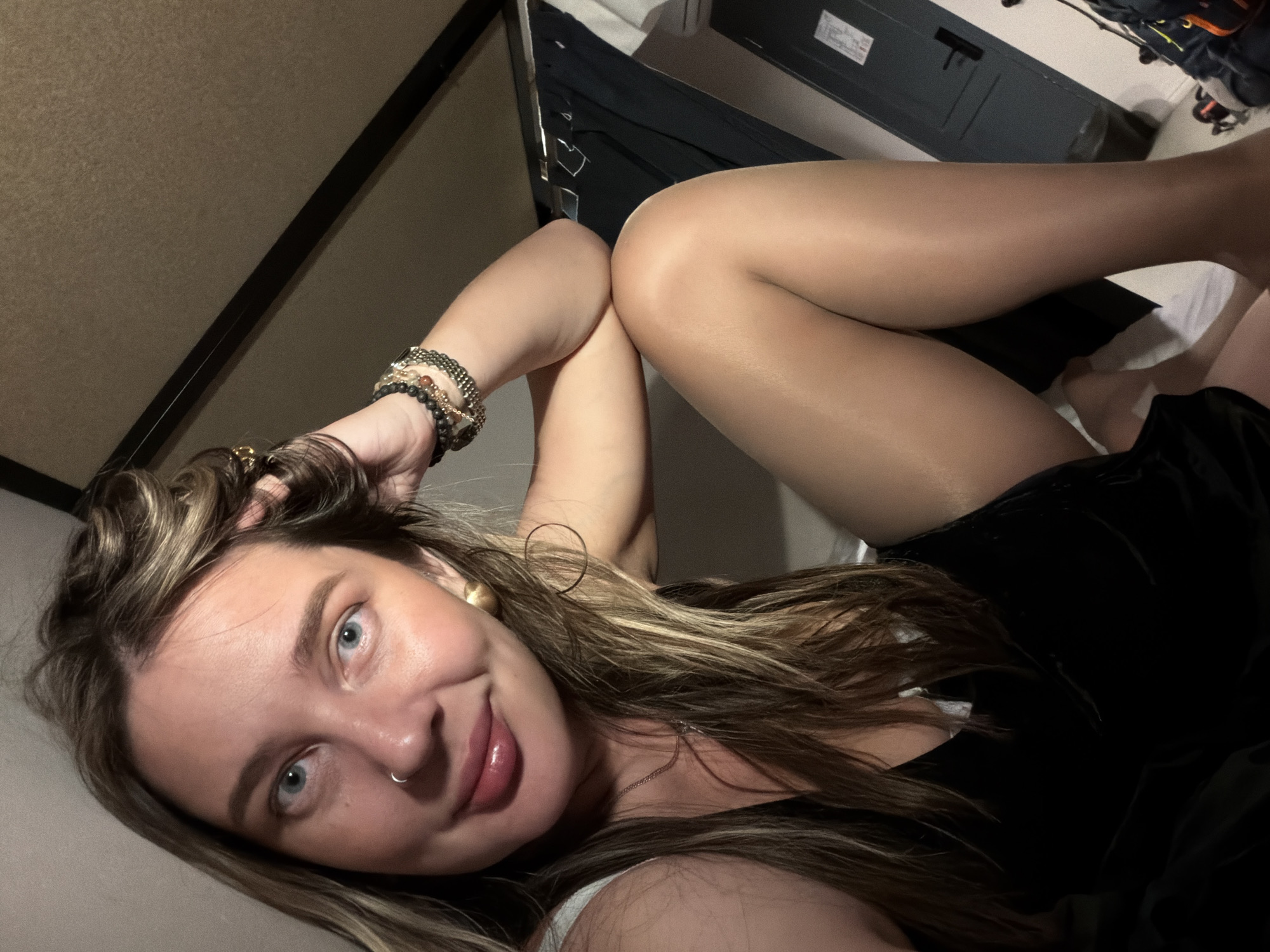 A young woman with long, highlighted brown hair is reclining on a bed. She is smiling at the camera with her head tilted to the left. She has light blue eyes and a silver nose ring. She is wearing a black sleeveless top and nude-colored pantyhose. Her arms are crossed above her knees, and she is wearing multiple beaded bracelets on her right wrist.
The background includes a metal bed frame, a luggage compartment overhead, and a white sheet on the bed. There is also luggage visible in the upper right corner. The location is not discernible as Moscow, Russia without additional information.