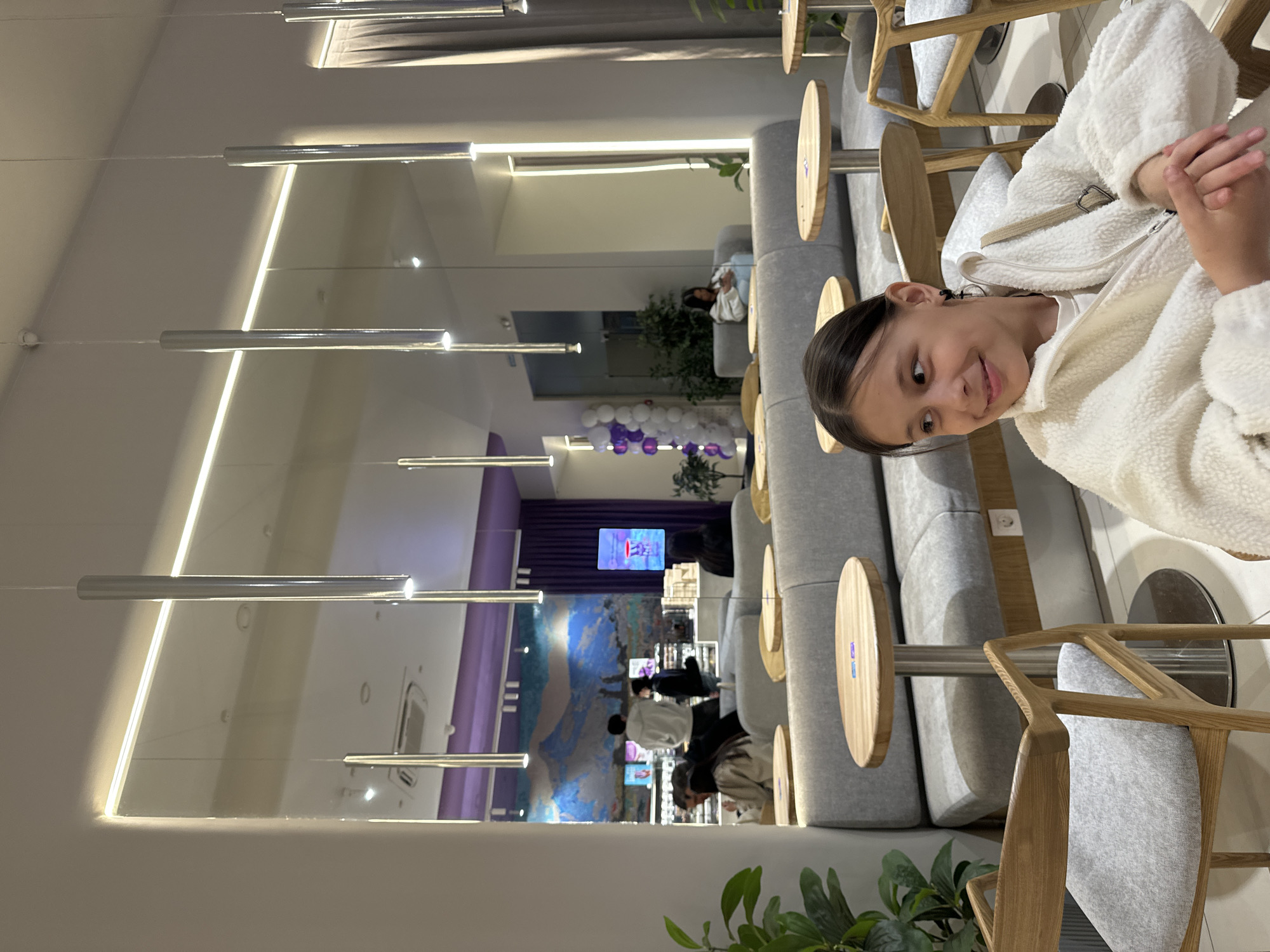 A vertical, medium shot shows a young girl sitting in an interior setting, possibly a lounge or waiting area, in the city of Unknown, Unknown. The girl is smiling towards the camera, wearing a white fluffy jacket over a white t-shirt, with her dark hair neatly pulled back. She is seated on a gray chair with a wooden frame. A small, round wooden table is affixed to the side of her seat.
The environment around the girl features modern design elements. In the background, a long, curved gray couch stretches along the wall. On the wall behind the couch, there's a mural-like design, possibly depicting a landscape or abstract art with hues of blue and purple. A small television screen is visible on the wall in the distance.
Overhead, long, cylindrical lights hang from the ceiling, providing a modern and stylish illumination to the space. In the distance, other people can be seen seated or moving about, suggesting this is a public or semi-public space. The overall color palette is subdued, with grays, whites, and soft blues dominating the scene. A potted green plant adds a touch of nature to the interior.