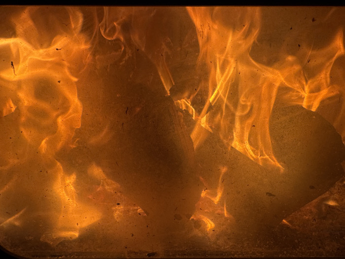The image depicts a close-up, internal view of an active combustion process occurring within an enclosed chamber. Prominent are large, dynamic flames, predominantly colored in vivid orange and yellow hues, with undertones of red and amber, indicating intense heat. These flames undulate and rise, occupying the majority of the frame. Embedded within and partially obscured by the flames are multiple irregularly shaped, dark solid objects, consistent with pieces of combustible material, such as wood logs, undergoing active burning.
The scene is viewed through a translucent barrier, likely a glass panel, which exhibits a scattered distribution of small, dark particulate matter, identified as soot, ash, or dust, accumulated on its surface. A narrow, dark, solid border is visible along the top edge of the image, with a thinner similar line at the bottom, suggesting the structural framing of the combustion chamber's viewing window. No people, specific actions, or interactions are discernible. The precise location within the city of Profesor Ishirkovo, Bulgaria, is not discernible from the visual evidence presented.
