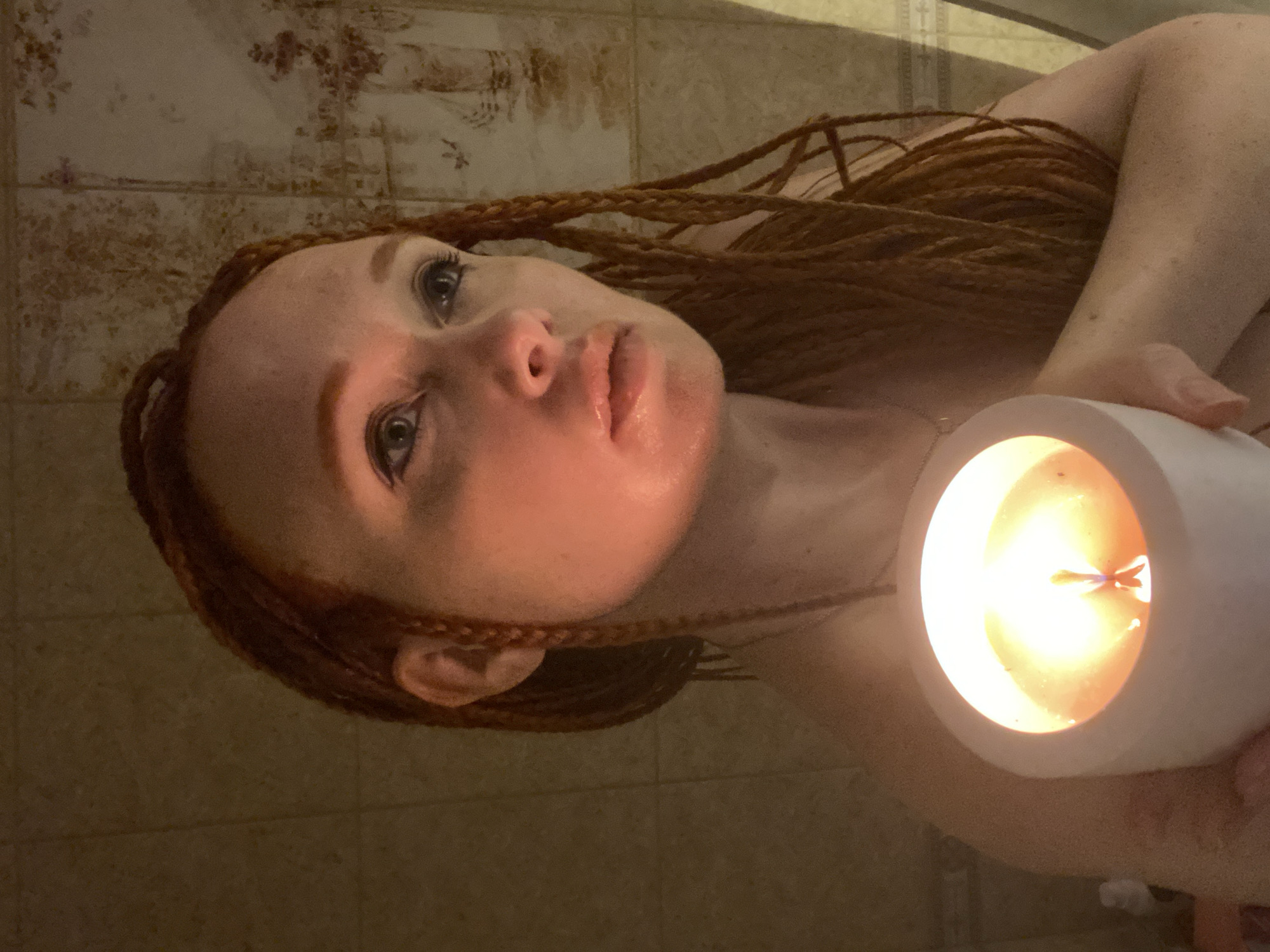 A close-up shot shows a woman with pale skin, freckles, and long, braided red hair. She is holding a lit white candle in a cylindrical holder with her right hand. The candle flame illuminates her face, casting a warm glow. She is looking off to her right with her lips slightly parted. She is wearing a necklace with a small pendant. The background consists of tiled walls, with a decorative pattern visible on the right side, possibly in a bathroom or spa setting. The lighting is dim and atmospheric. There is no indication of the city of Sochi, Russia, in the image.