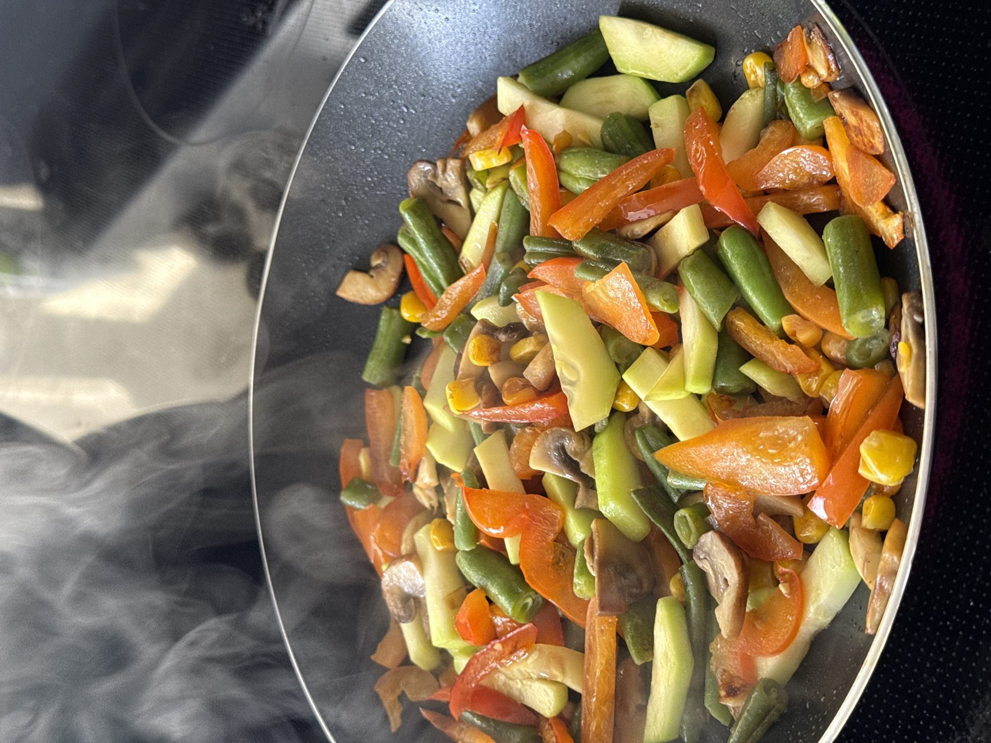 A close-up, overhead shot shows a variety of chopped vegetables being stir-fried in a black non-stick skillet. The vegetables include sliced green beans, diced red bell peppers, sliced zucchini, sliced mushrooms, and corn kernels. Steam rises from the skillet, indicating the food is hot. The skillet is on a black stovetop. The background is out of focus, but appears to be a kitchen environment. There is no indication of the city of Chicago, United States of America.